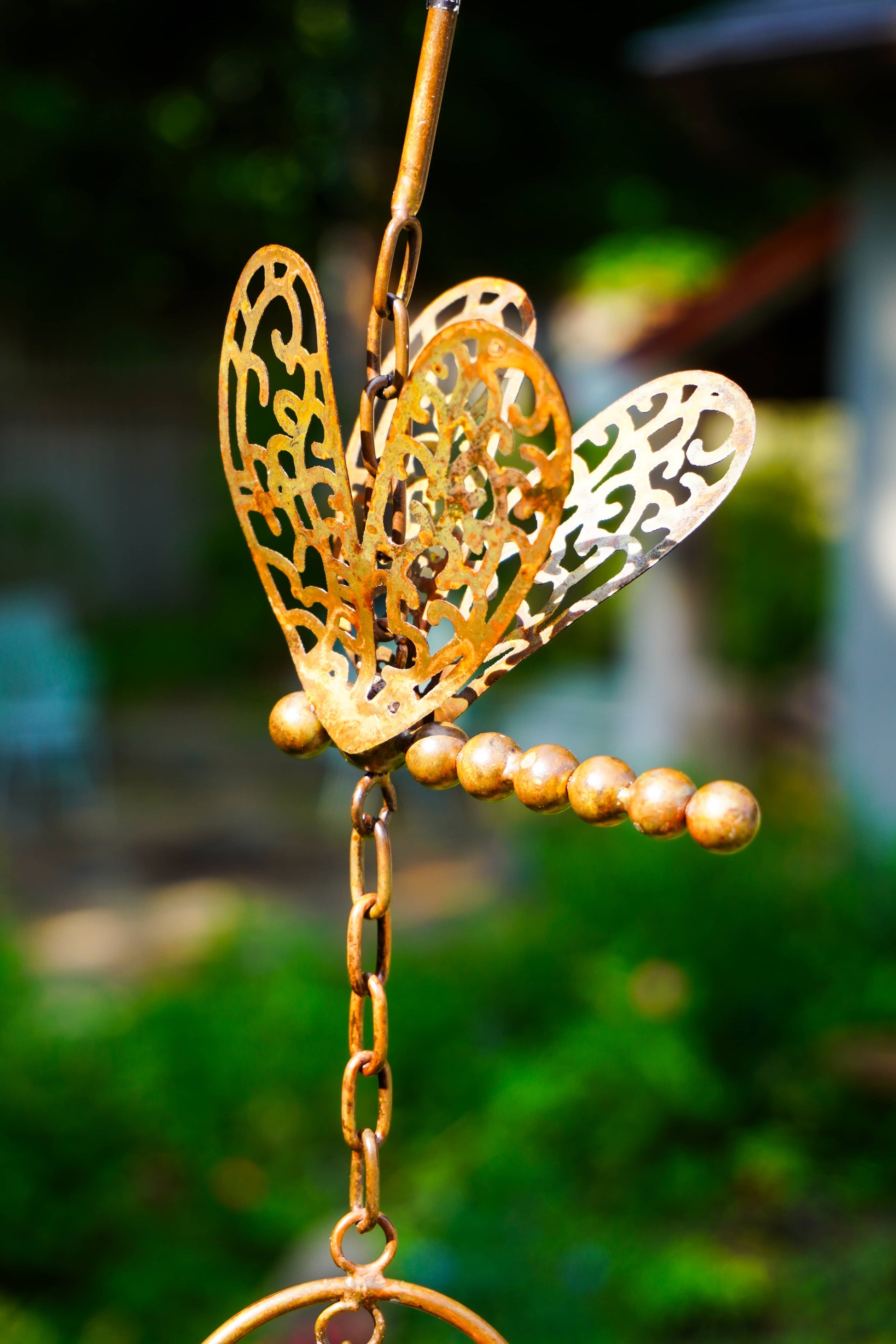A brass dragonfly garden ornament with intricate cutout wings and a beaded stem, shown hanging outdoors against a blurred green garden background.