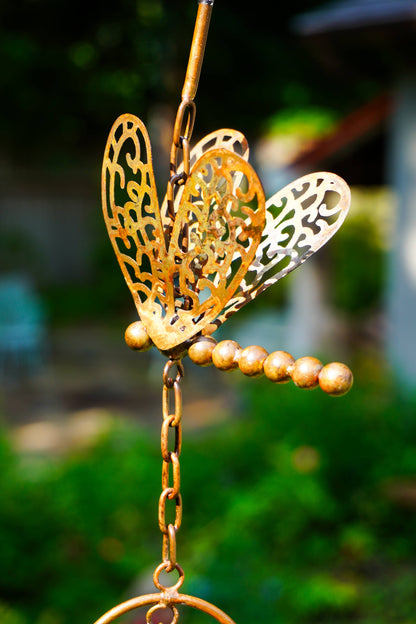 A brass dragonfly garden ornament with intricate cutout wings and a beaded stem, shown hanging outdoors against a blurred green garden background.