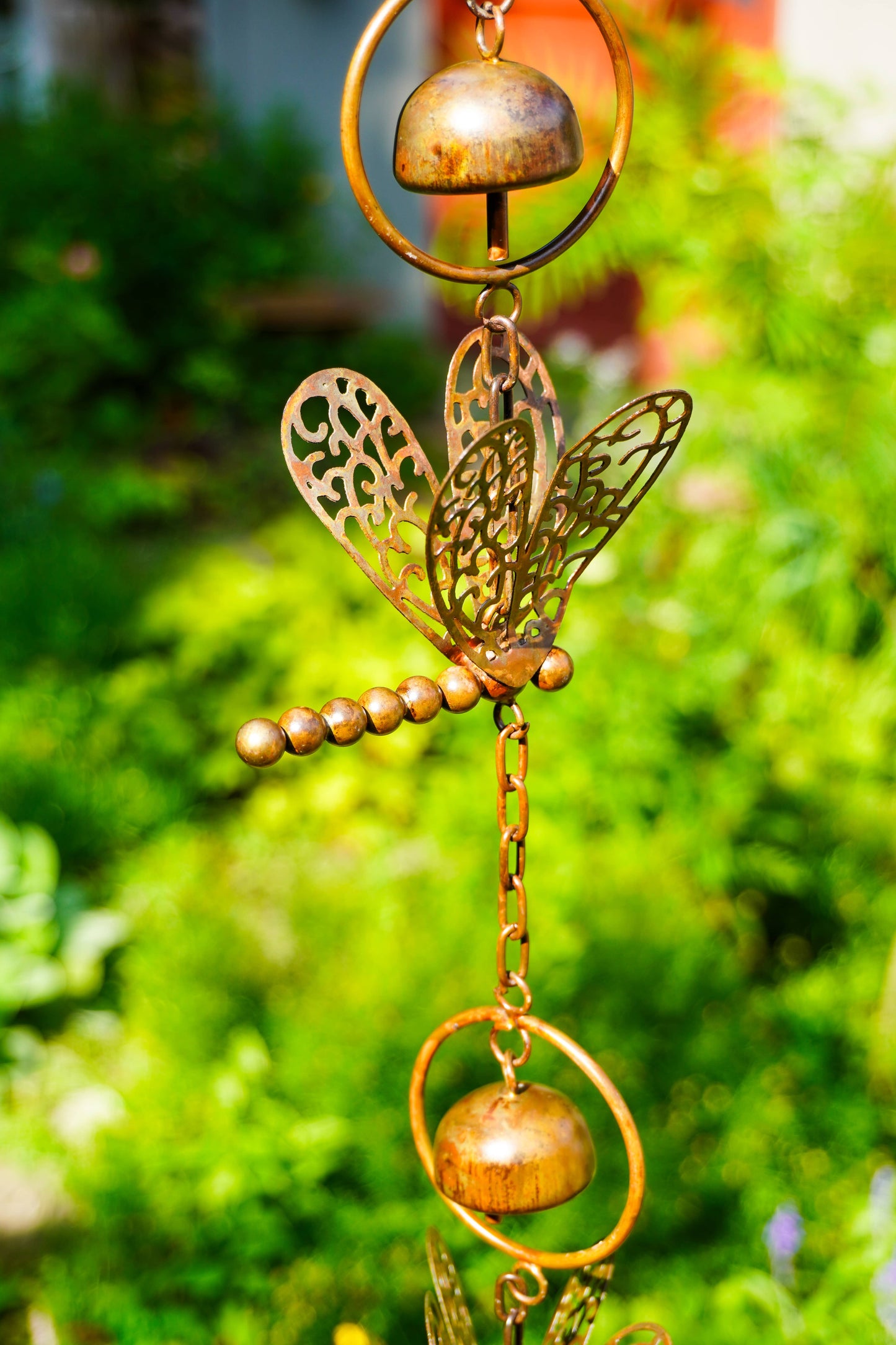 A bronze metal garden wind chime featuring a dragonfly, beaded stem, and hanging bells, shown against a blurred green garden background.