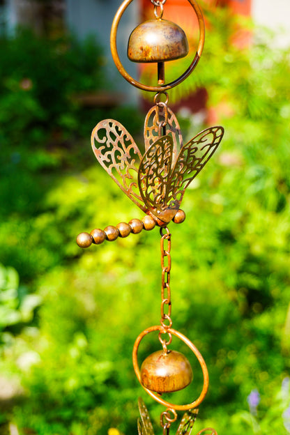 A bronze metal garden wind chime featuring a dragonfly, beaded stem, and hanging bells, shown against a blurred green garden background.