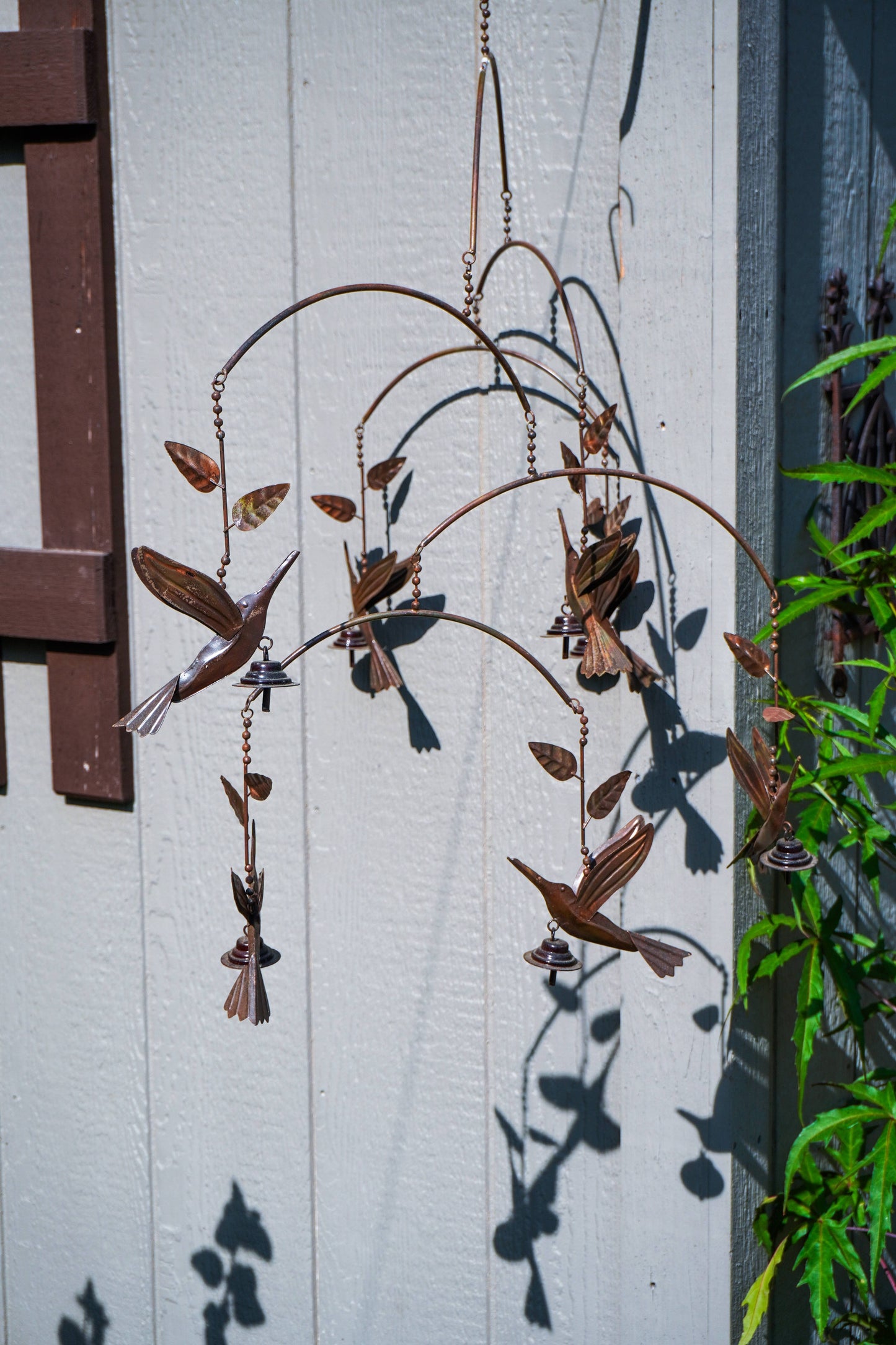 A metal hummingbird garden wind chime hanging against a white paneled wall, with green plants visible on the right side and strong sunlight casting shadows.