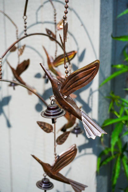 Metallic bronze hummingbird mobile hanging outdoors against a light gray wall, with leaf-shaped branches and small wind chimes.