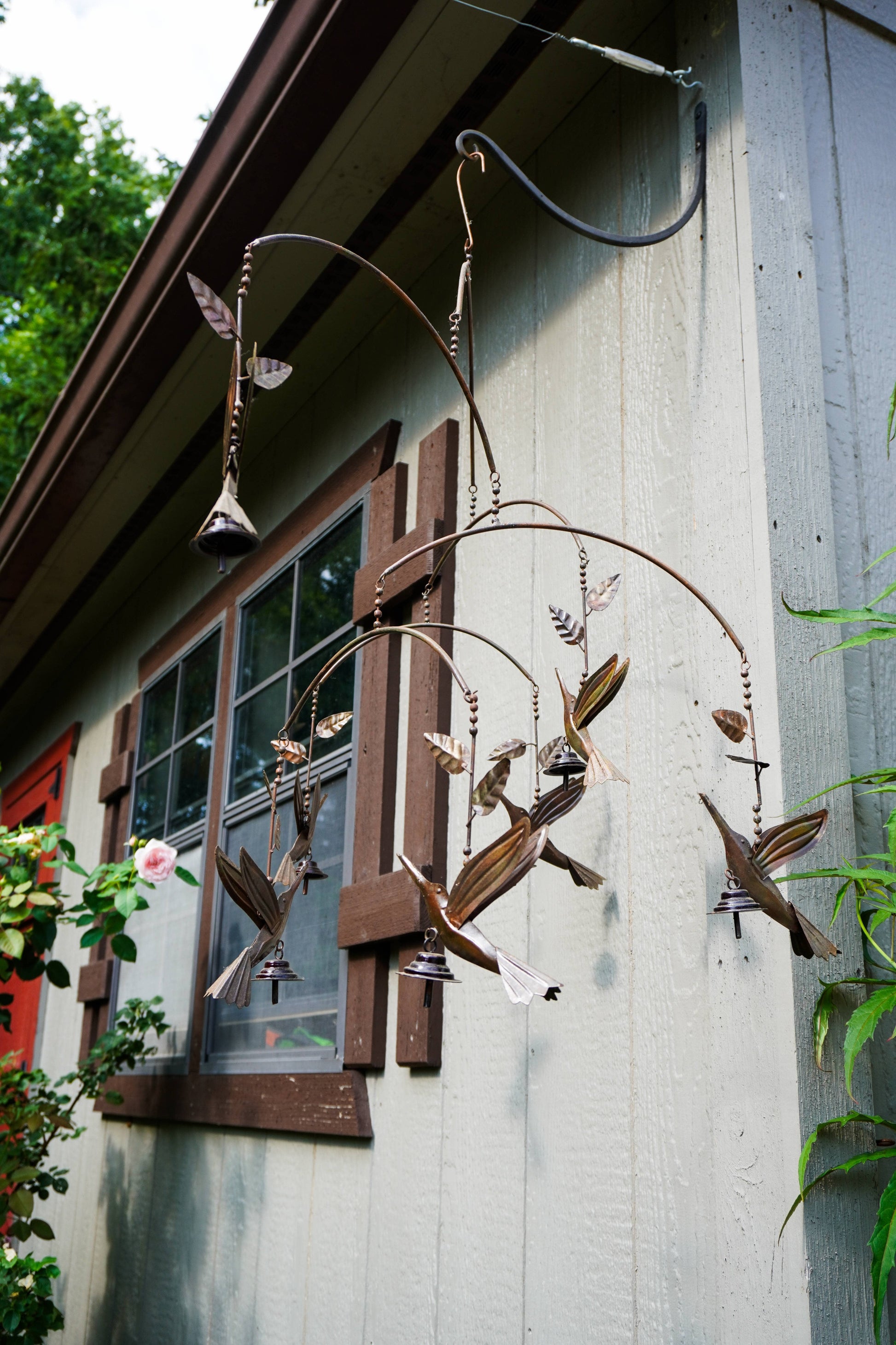 A metal hummingbird wind chime with small bells and hanging branches, mounted on the exterior wall of a house next to a window.