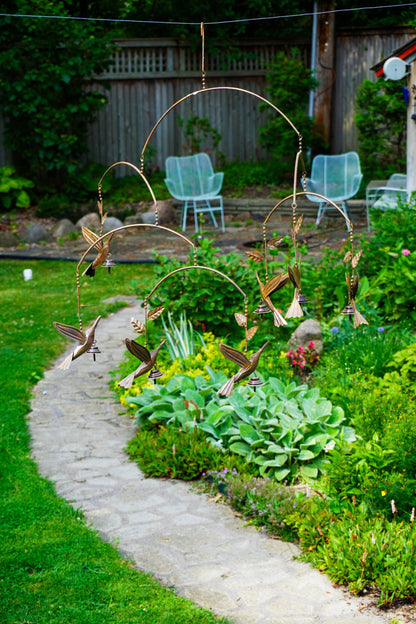 A garden mobile with hanging birds and wheat motifs suspended from curved metal rods, displayed outdoors in a landscaped garden next to a stone path.