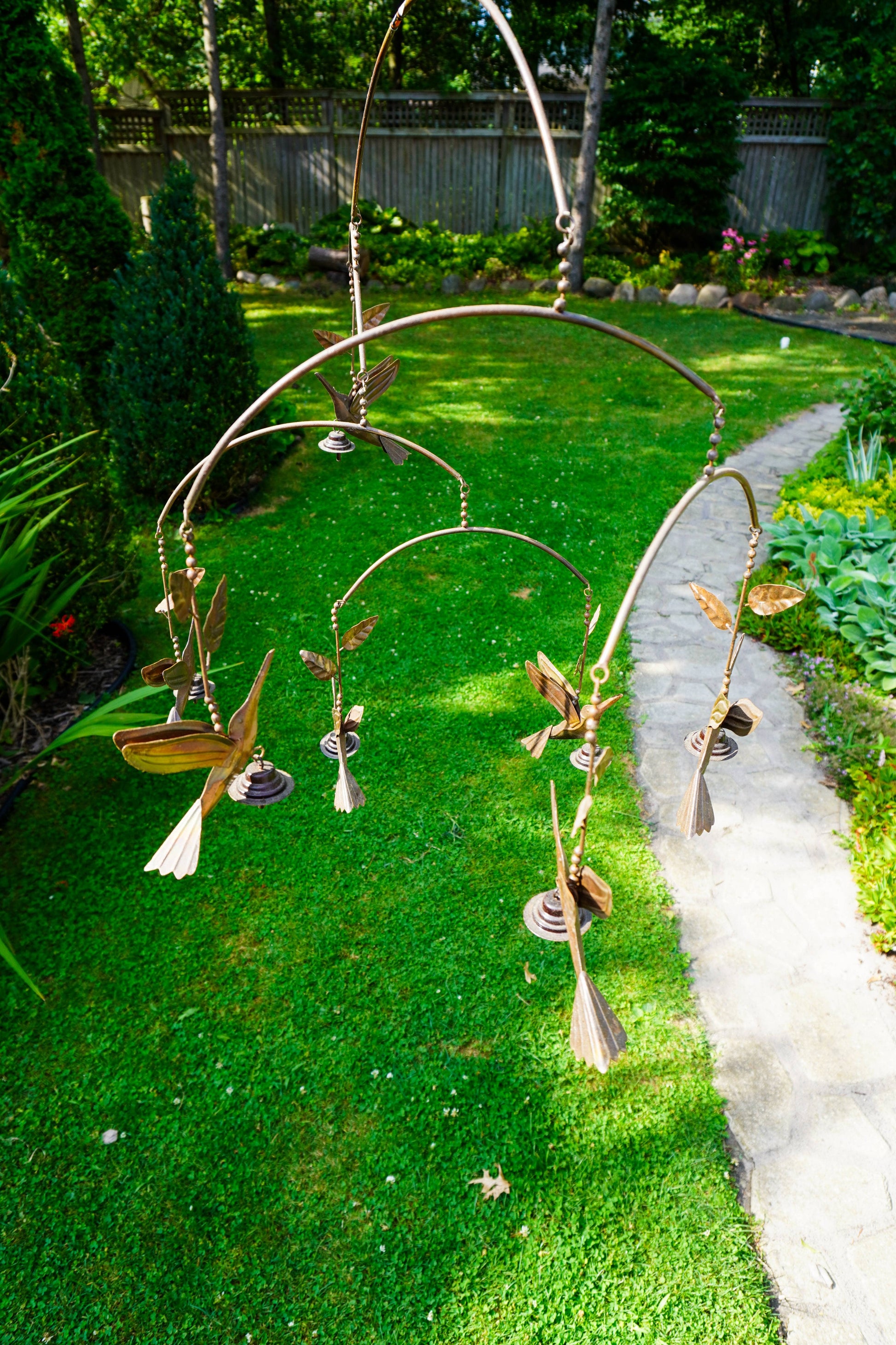 A metal bird mobile hanging outdoors in a garden, featuring several hanging birds and leaves suspended from curved metal rods over a green lawn and concrete path.