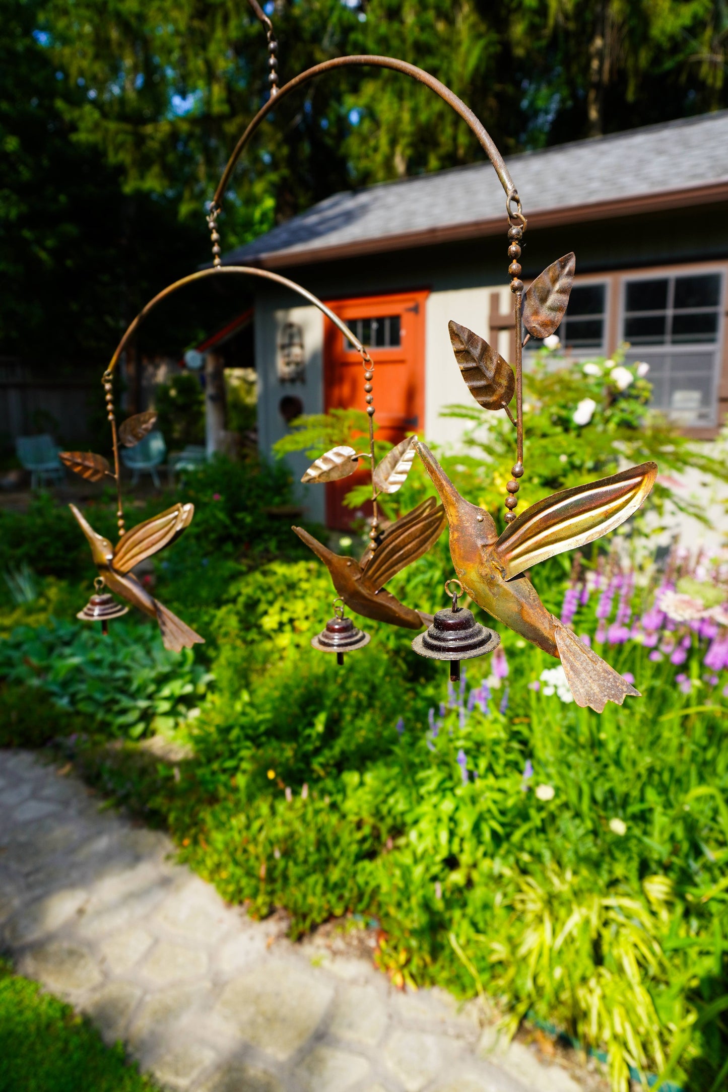 Metal bird mobile hanging in a lush garden with green plants, a stone path, and a house in the background.