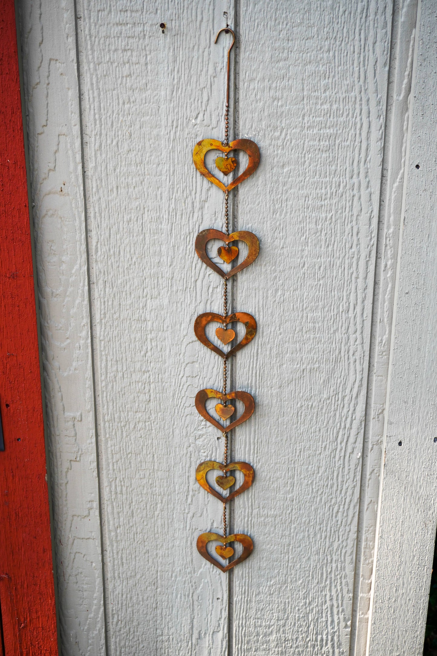 A rustic metal heart wind spinner hanging vertically against a white painted wooden door. The ornament features seven stacked, concentric cutout hearts in a weathered rust-orange and brown finish, suspended from a thin chain with a hook at the top.