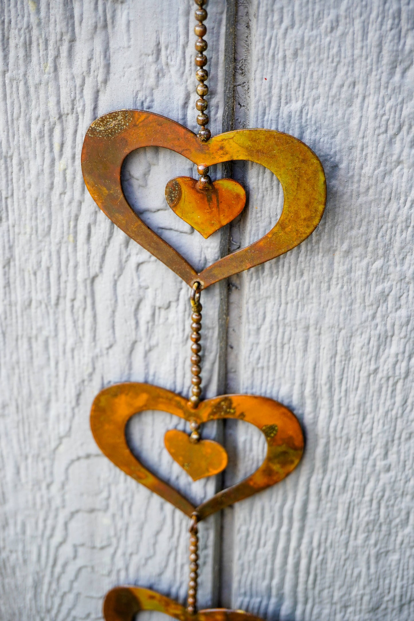 A rustic metal heart-shaped hanging decoration with a small amber glass heart inside each larger metal heart, suspended on a beaded chain against a white textured wooden background.