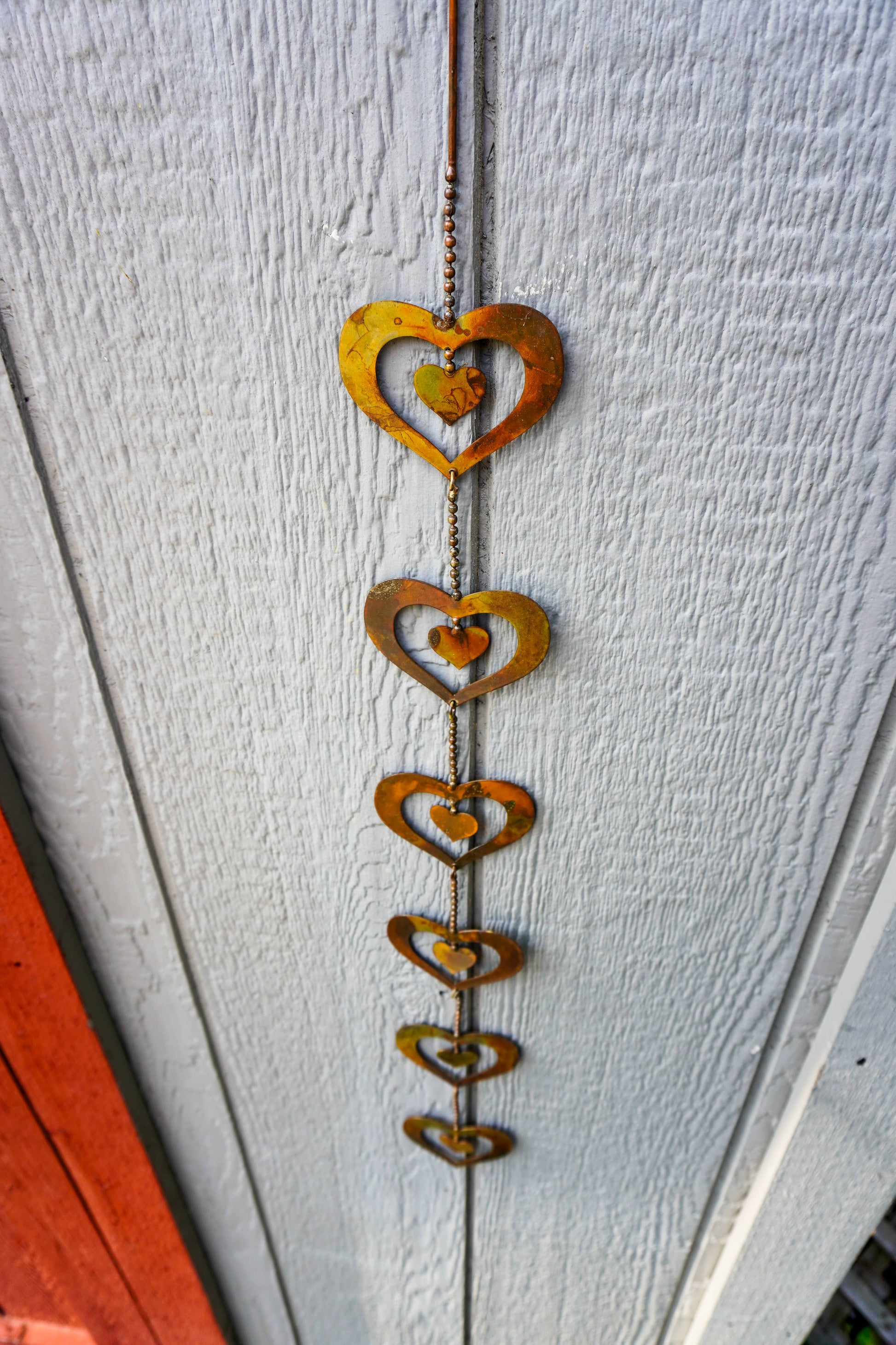 A rustic metal heart wind spinner hanging vertically against a white painted wooden door. The decorative wind spinner features a series of nested, cutout hearts in varying sizes, finished with a weathered rust-brown patina.