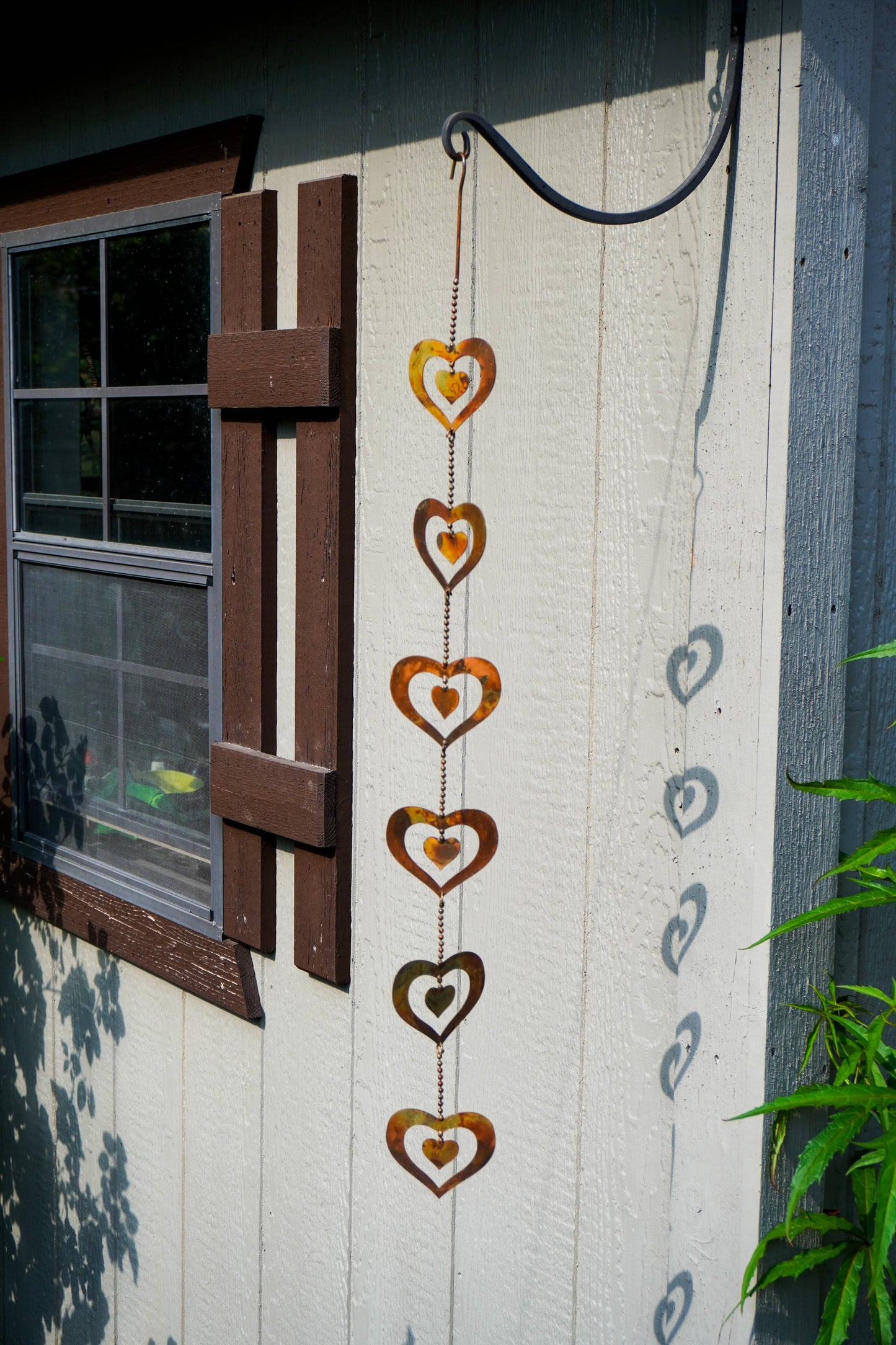 A vertical hanging metal heart wind spinner decoration with five stacked rusty brown hearts, shown hanging from a black hook on a white wooden fence next to a window.