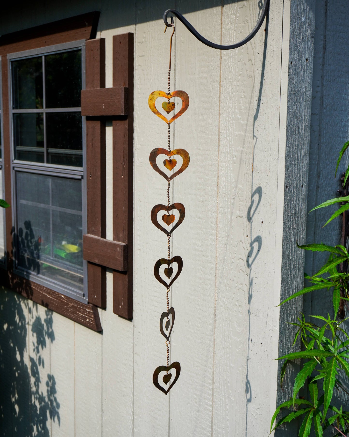 A vertical hanging metal heart ornament with five stacked cutout hearts in a rustic bronze finish, suspended outdoors against a white wood-paneled wall next to a window.