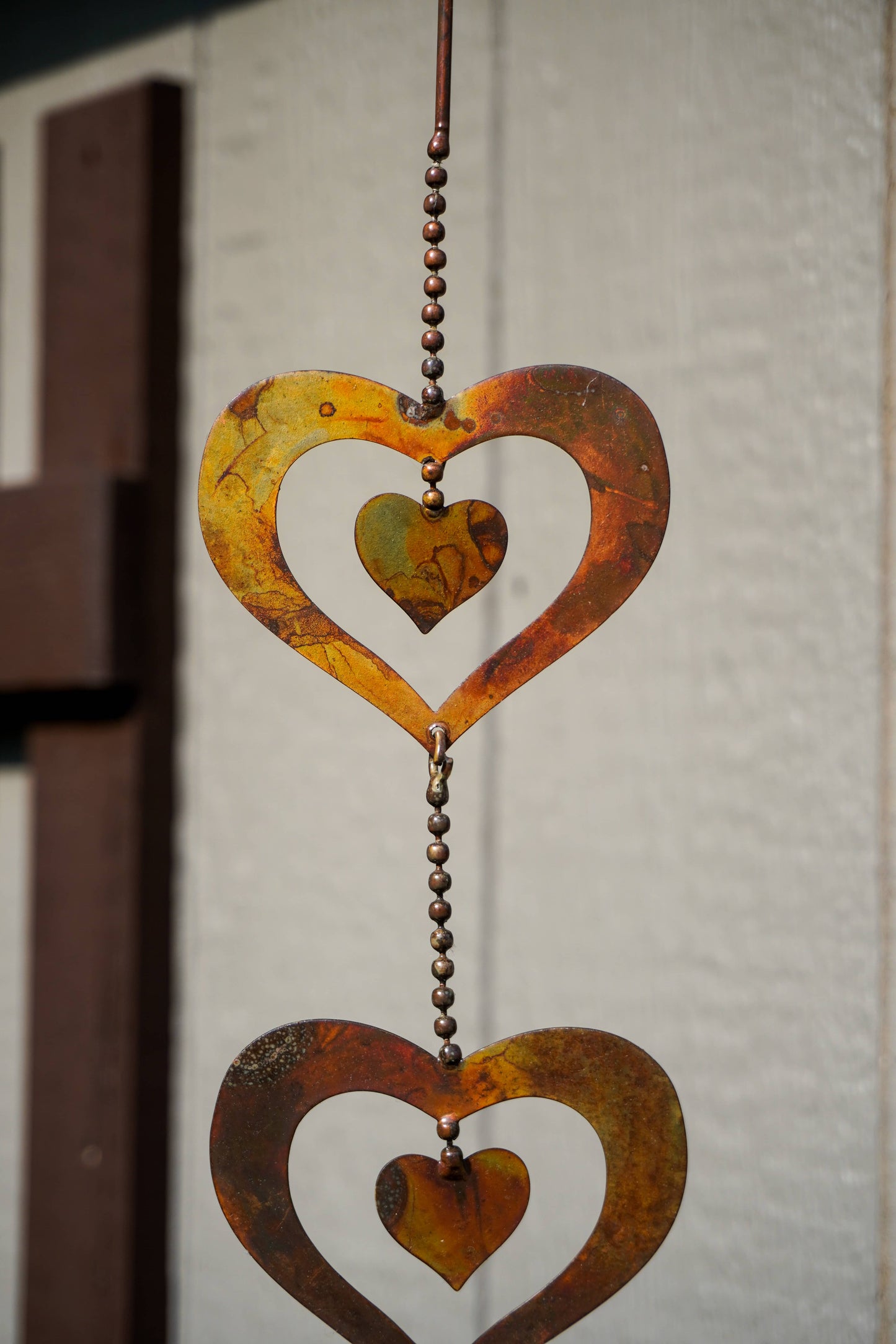 A rustic metal heart-shaped hanging ornament with a textured rust finish, suspended by a beaded chain against a neutral textured wall.