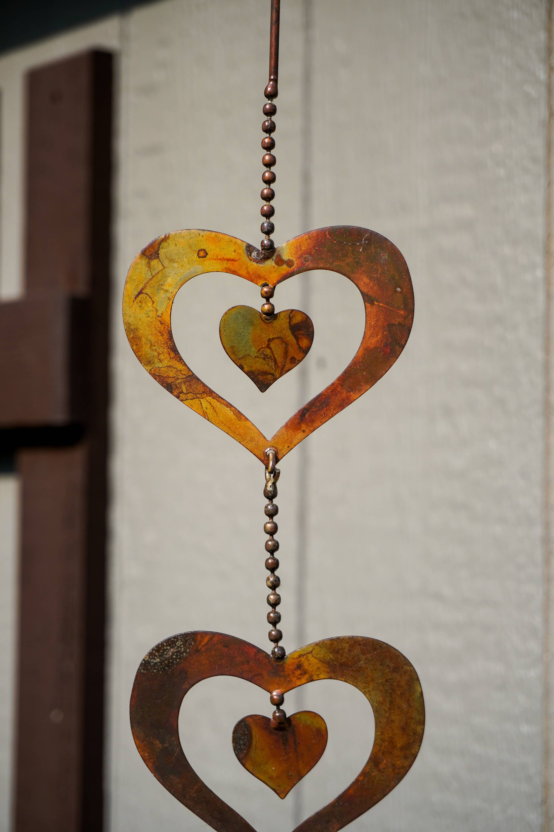 A rustic metal heart-shaped hanging ornament with a textured rust finish, suspended by a beaded chain against a neutral textured wall.