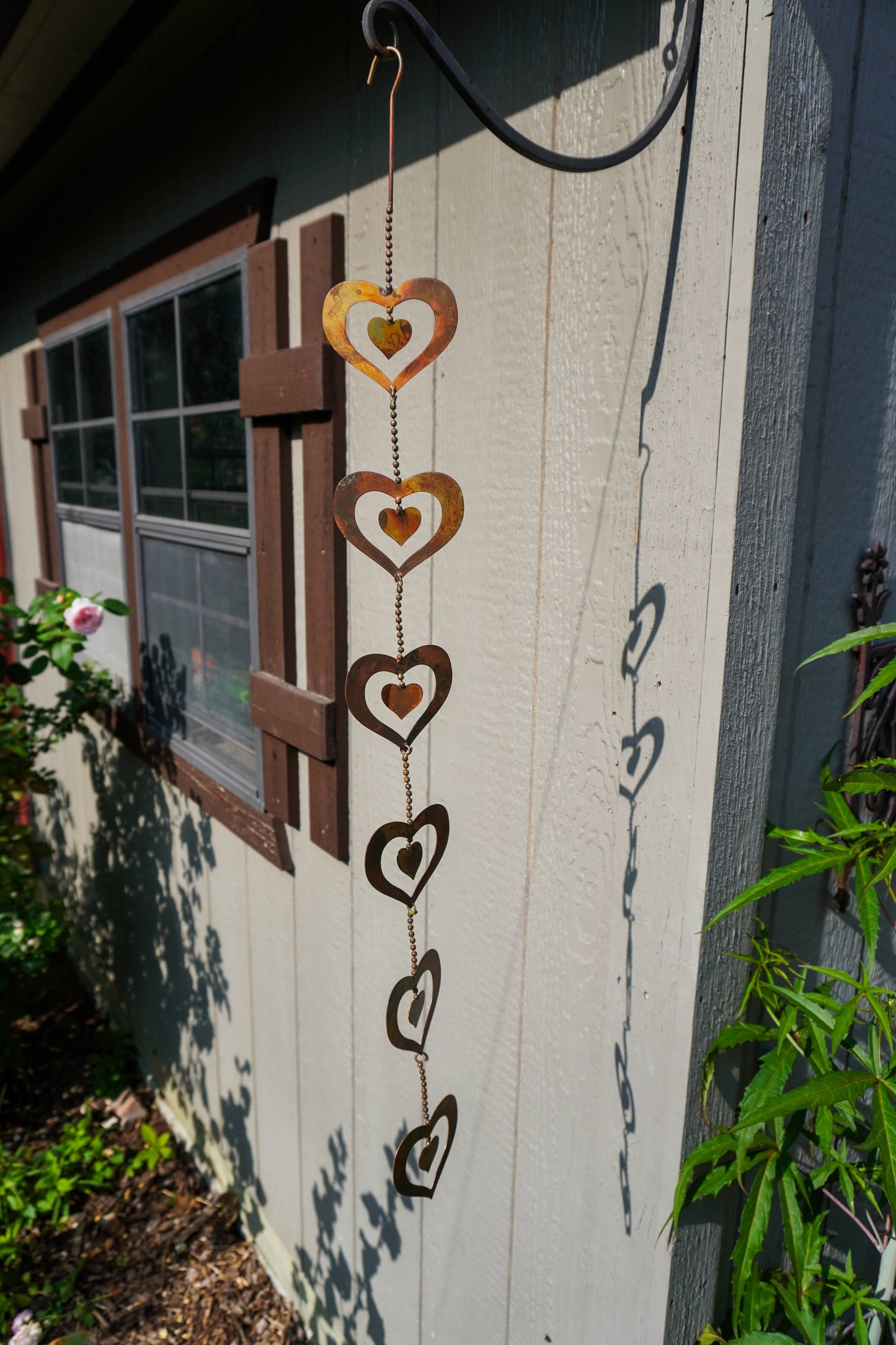 A rustic metal heart wind spinner hanging outdoors against a light gray wooden house wall next to a window.