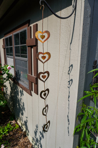 A rustic metal heart wind spinner hanging outdoors against a light gray wooden house wall next to a window.