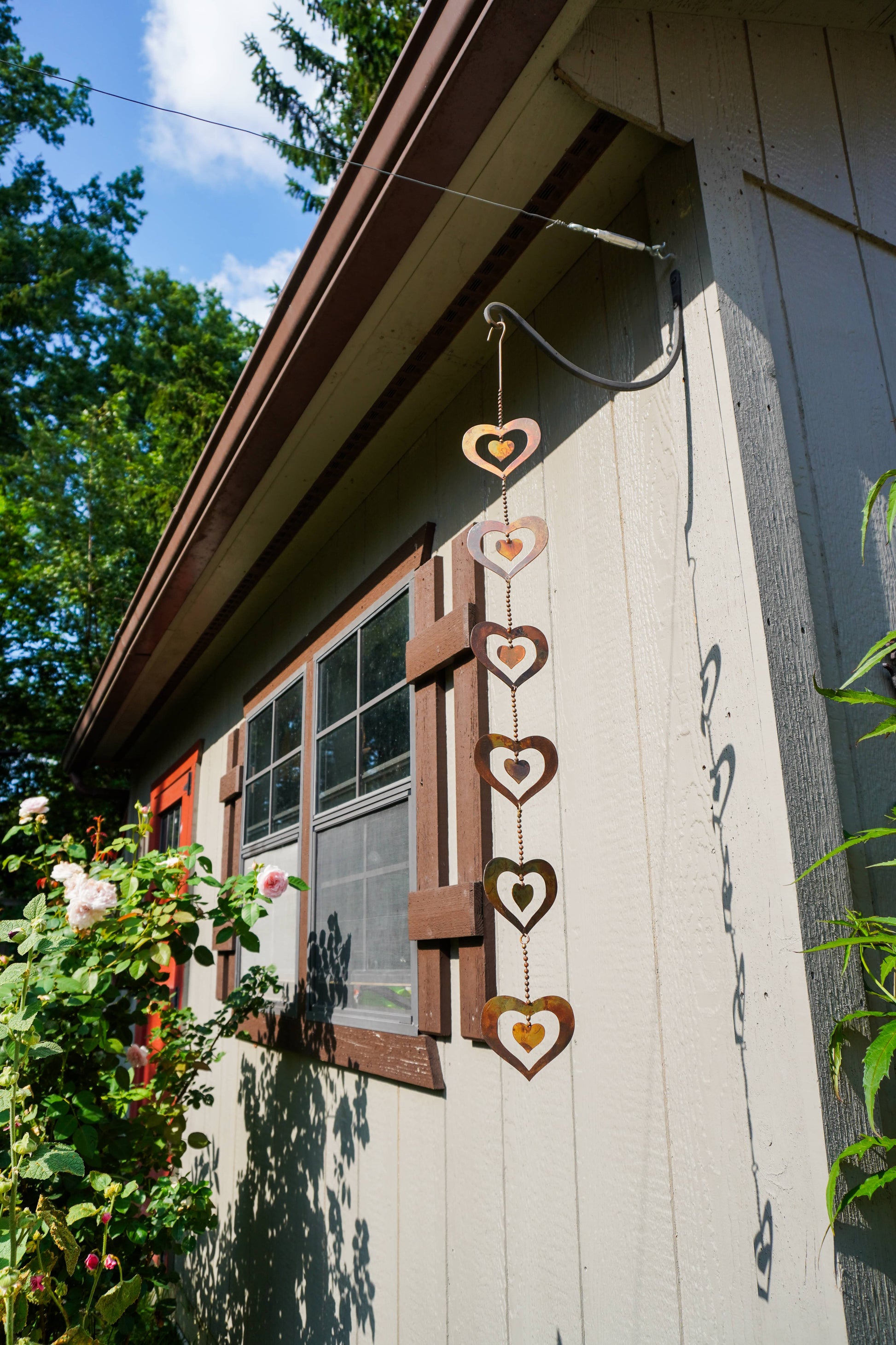 A vertical hanging metal heart wind spinner with five stacked heart shapes in rusted brown and warm gold, suspended outdoors against a house exterior wall with greenery and a window.