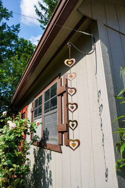 A vertical hanging metal heart wind spinner with five stacked heart shapes in rusted brown and warm gold, suspended outdoors against a house exterior wall with greenery and a window.