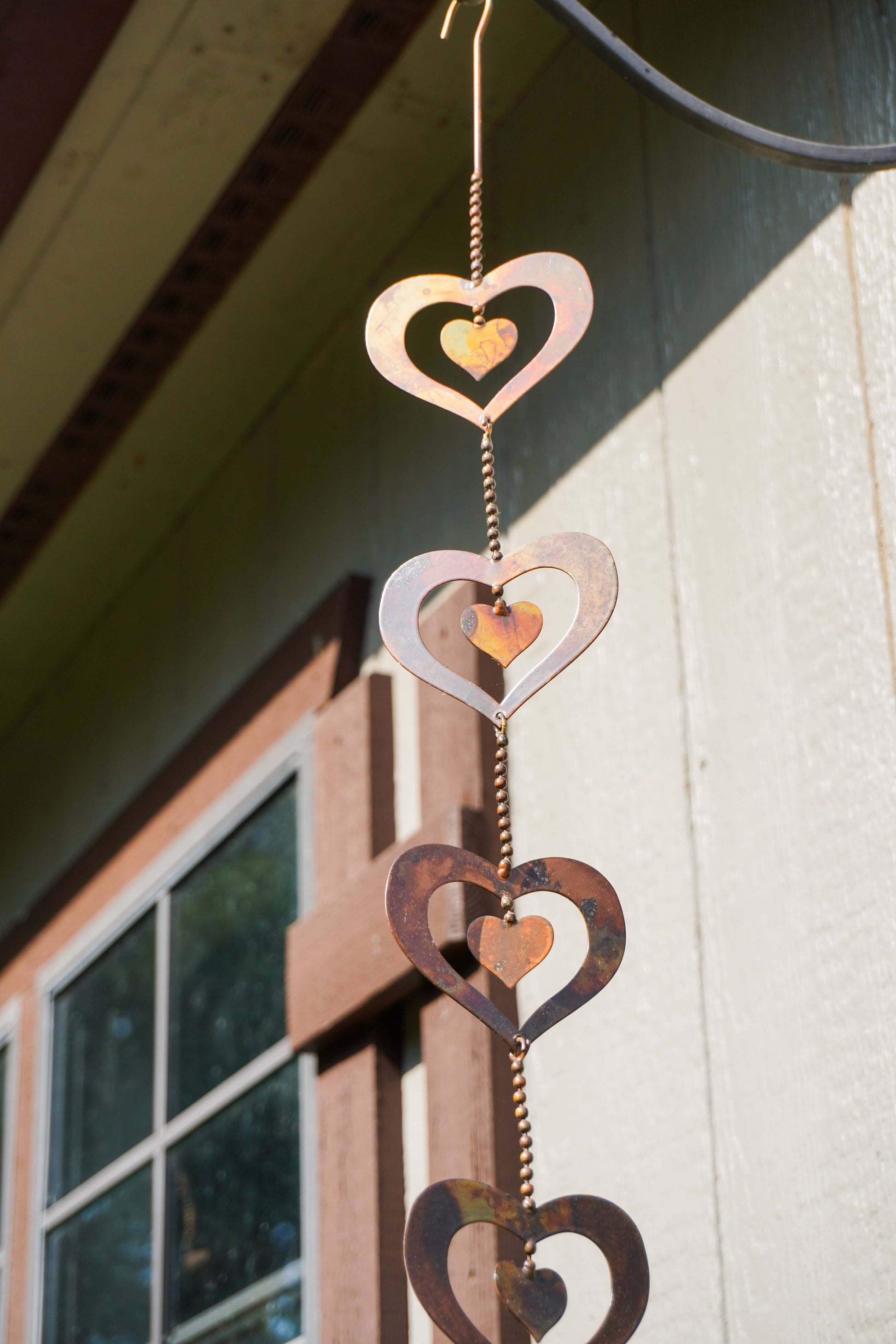 A hanging metal heart wind spinner with nested cutout hearts in copper, silver, and dark brown tones, photographed outdoors against a house wall and window.