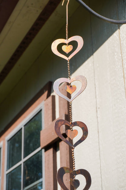 A hanging metal heart wind spinner with nested cutout hearts in copper, silver, and dark brown tones, photographed outdoors against a house wall and window.
