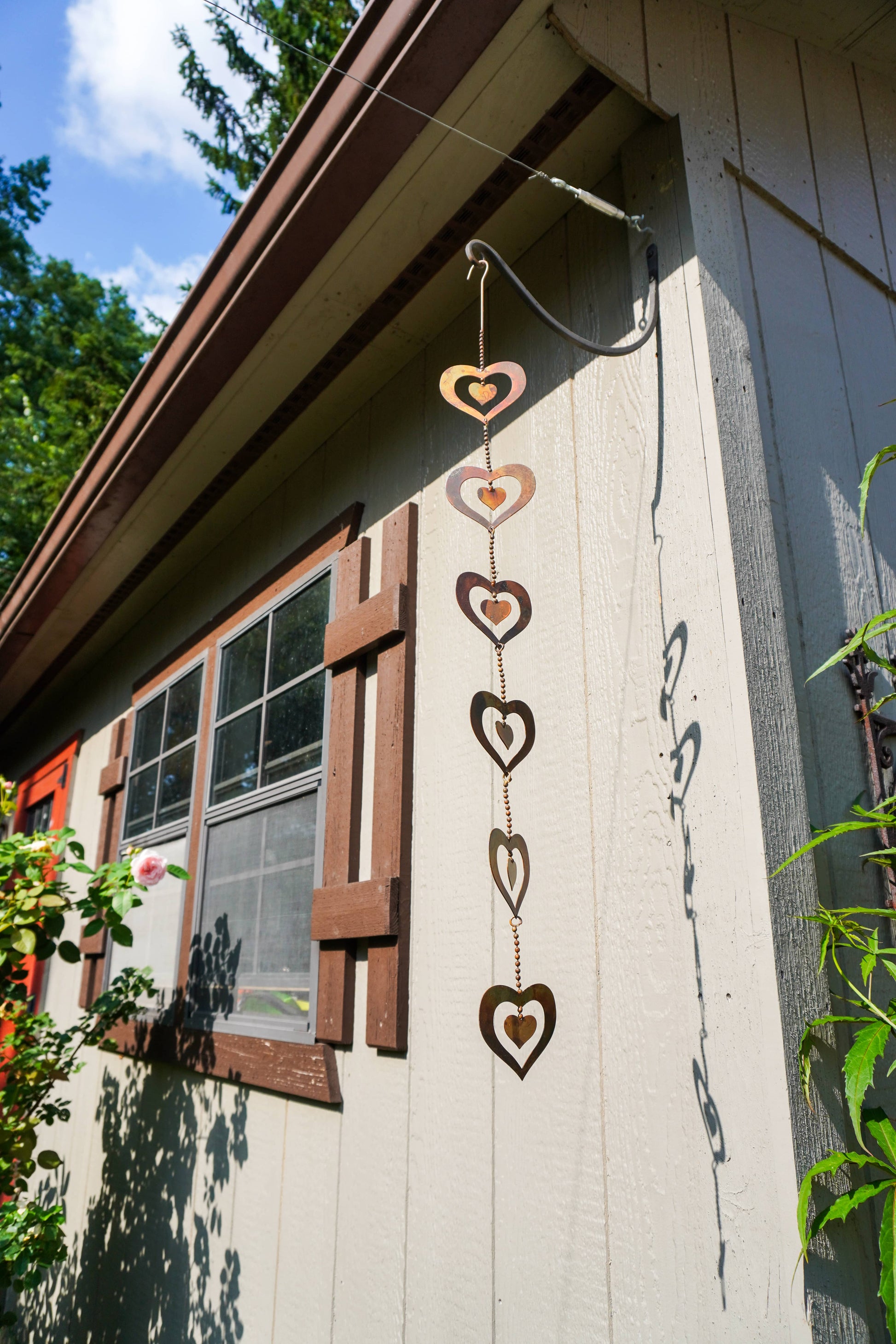 A vertical hanging metal heart wind spinner decoration with five nested heart shapes in varying sizes, shown hanging outside a house next to a window.