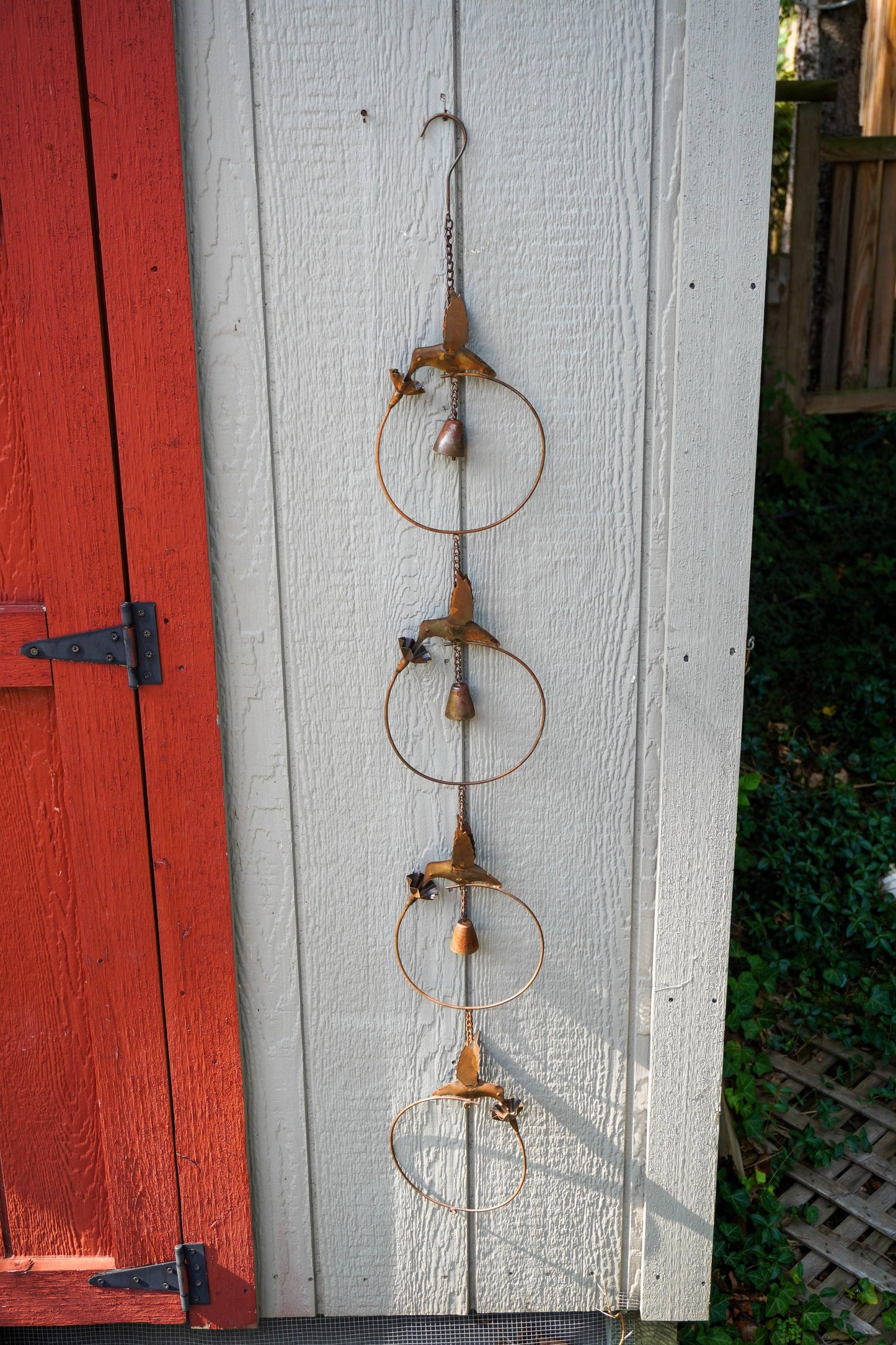 A rustic metal garden ornament hanging on a white wooden wall next to a red painted door, featuring three stacked circular rings with small wooden birds and beads.