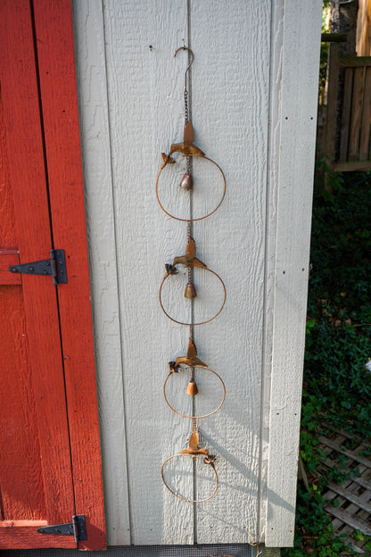 A rustic metal garden ornament hanging on a white wooden wall next to a red painted door, featuring three stacked circular rings with small wooden birds and beads.