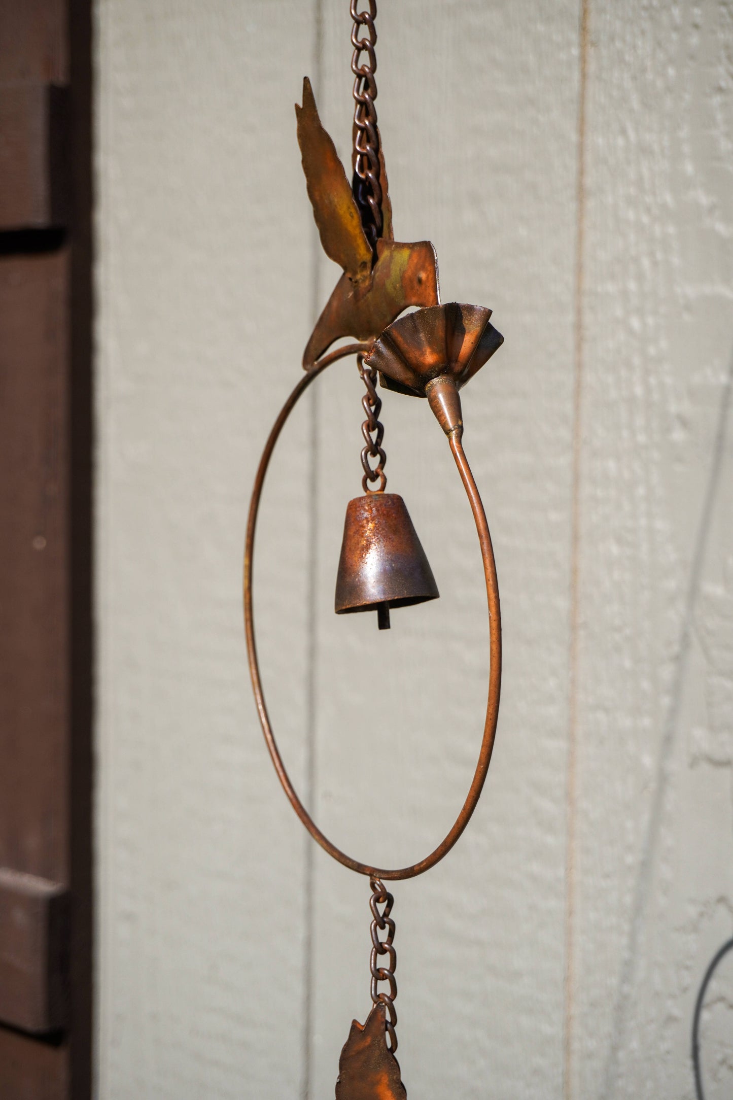 A copper metal wind chime featuring a hummingbird, flower, and bell suspended in a circular frame against a textured off-white wall.