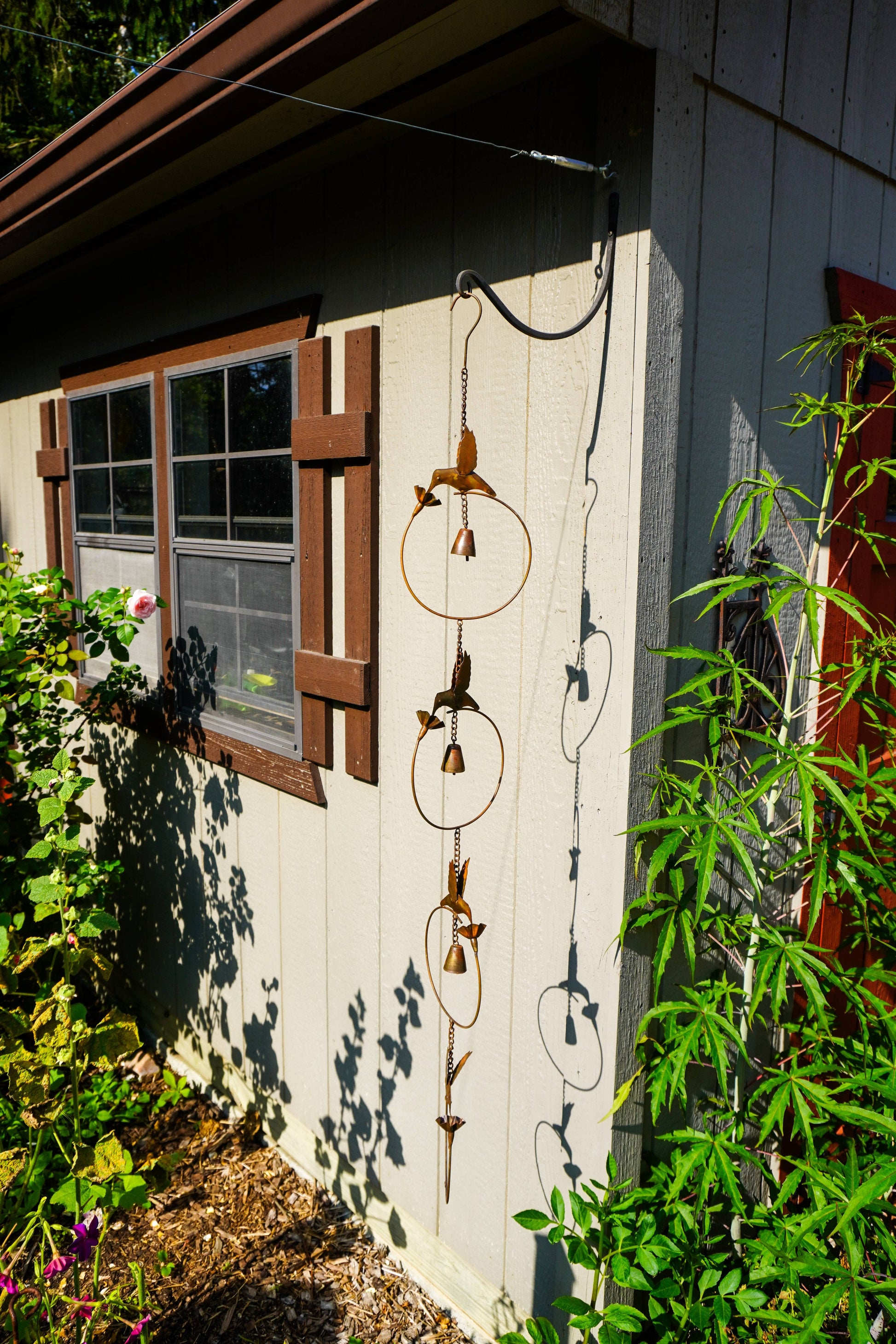 A rusted metal garden ornament with hanging rings and bird-like shapes hangs from a wire on the exterior wall of a house, surrounded by green plants and garden foliage in bright sunlight.
