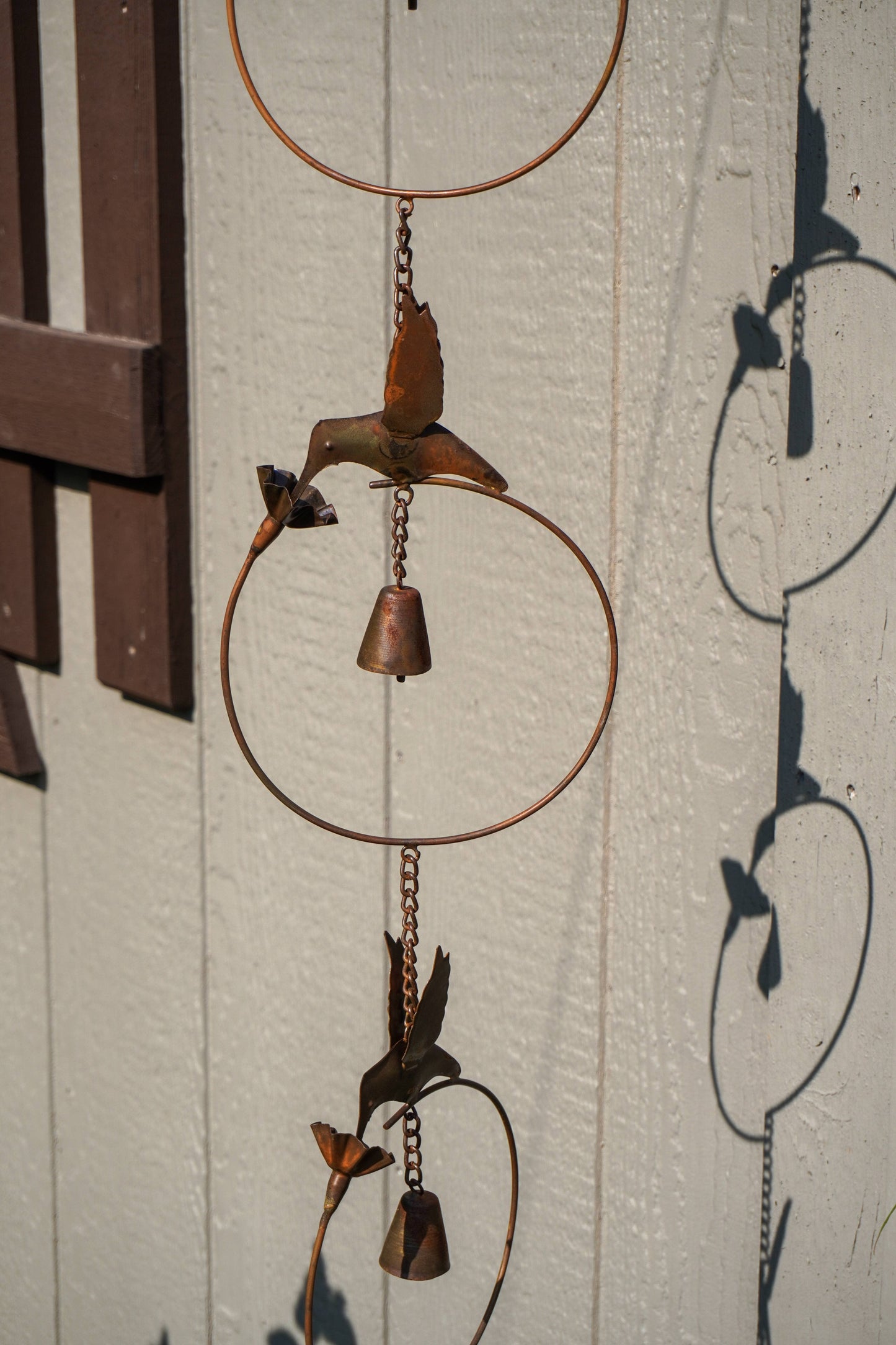 Rustic metal wind chime with circular hoops, hanging chains, and bird and flower shapes in a weathered copper-brown finish, photographed against a white paneled wall with strong sunlight and shadows.