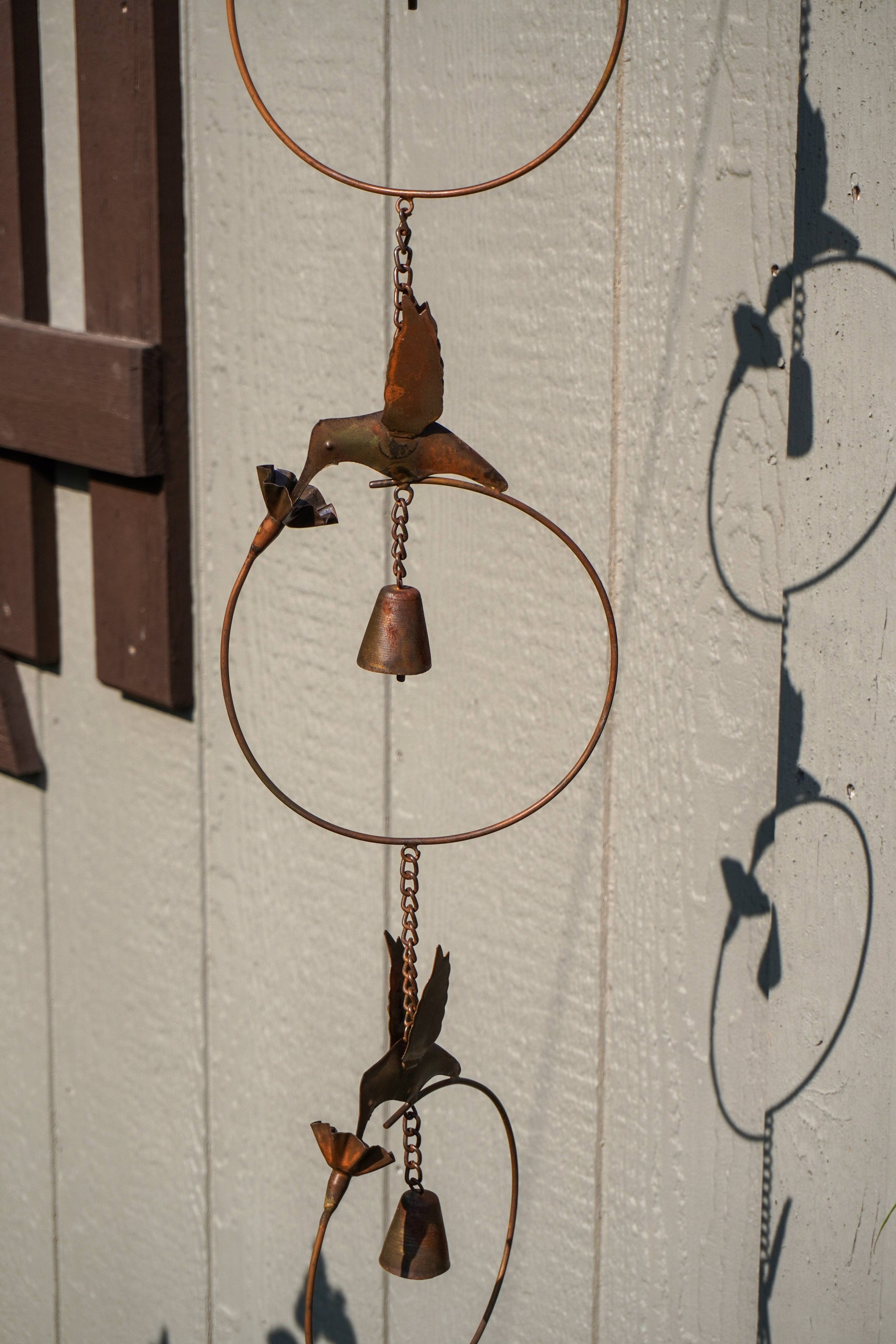 Rustic metal wind chime with circular hoops, hanging chains, and bird and flower shapes in a weathered copper-brown finish, photographed against a white paneled wall with strong sunlight and shadows.
