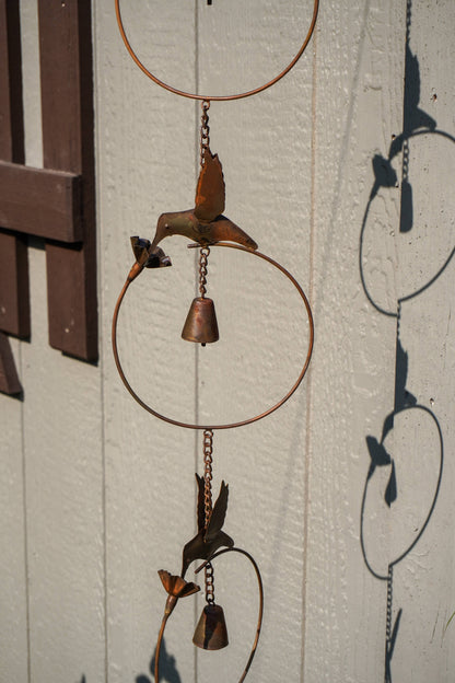 Rustic metal wind chime with circular hoops, hanging chains, and bird and flower shapes in a weathered copper-brown finish, photographed against a white paneled wall with strong sunlight and shadows.