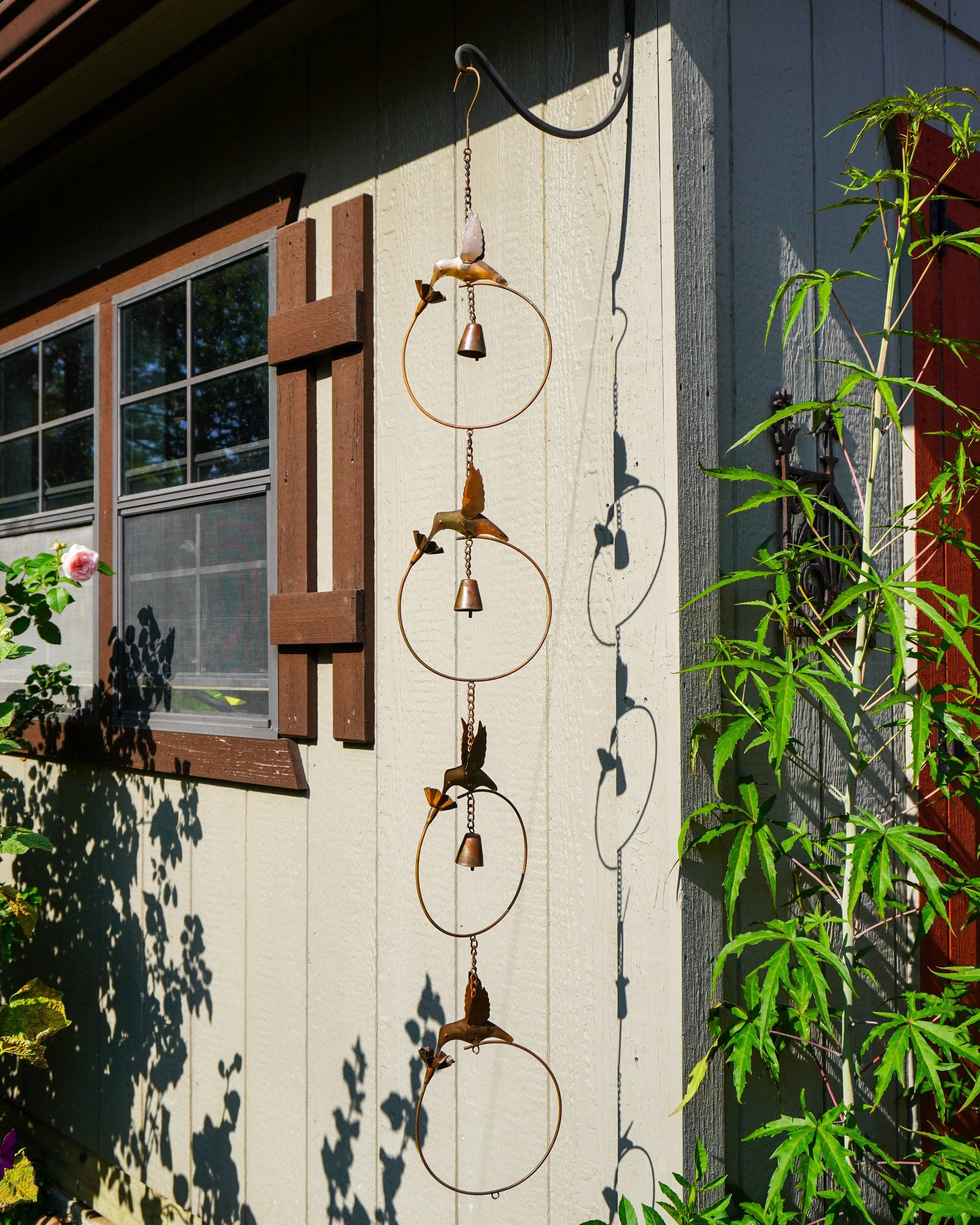 A rustic metal wind chime with bird and leaf motifs hangs outdoors against the side of a house. The hanging ornament features circular rings and small bird figures with dangling bells, set against a light gray siding wall with green plants and shadows.