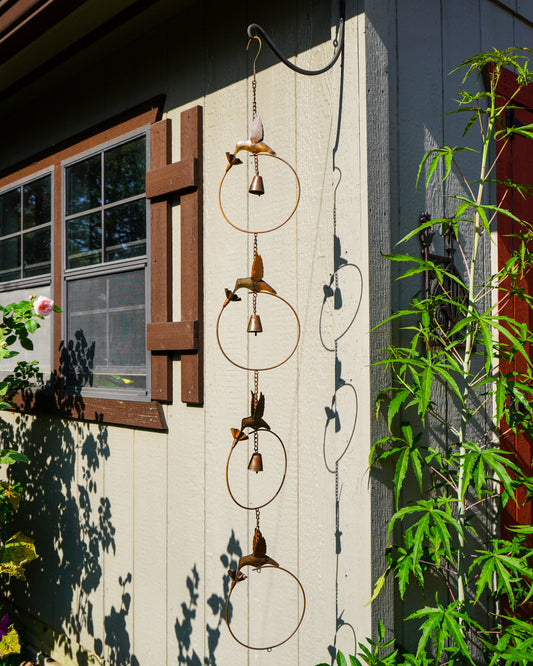 A rustic metal wind chime with bird and leaf motifs hangs outdoors against the side of a house. The hanging ornament features circular rings and small bird figures with dangling bells, set against a light gray siding wall with green plants and shadows.