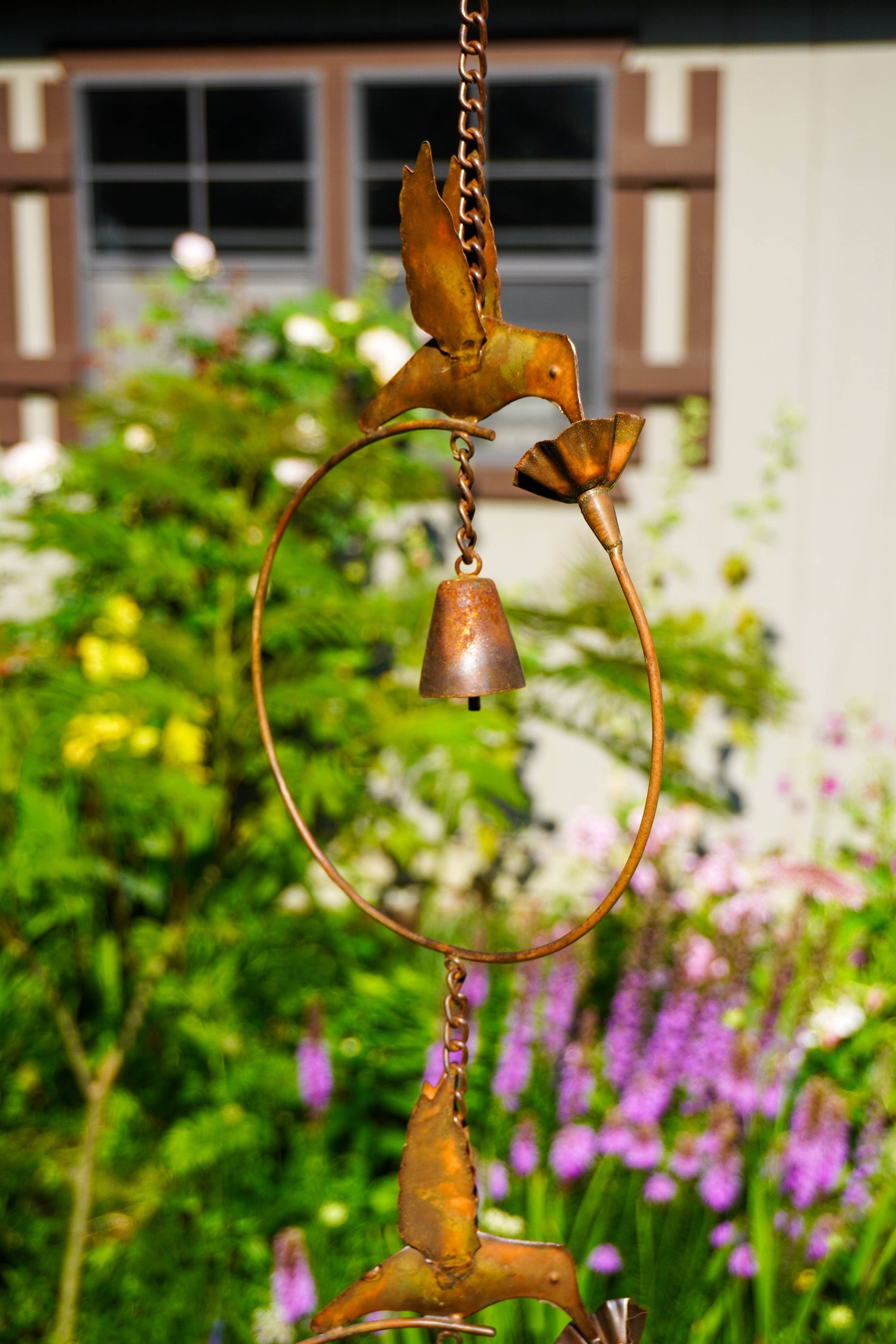 A rustic metal wind spinner hanging in a garden, featuring bird figures, a circular hoop, and a small bell, with a blurred background of green foliage and purple flowers.
