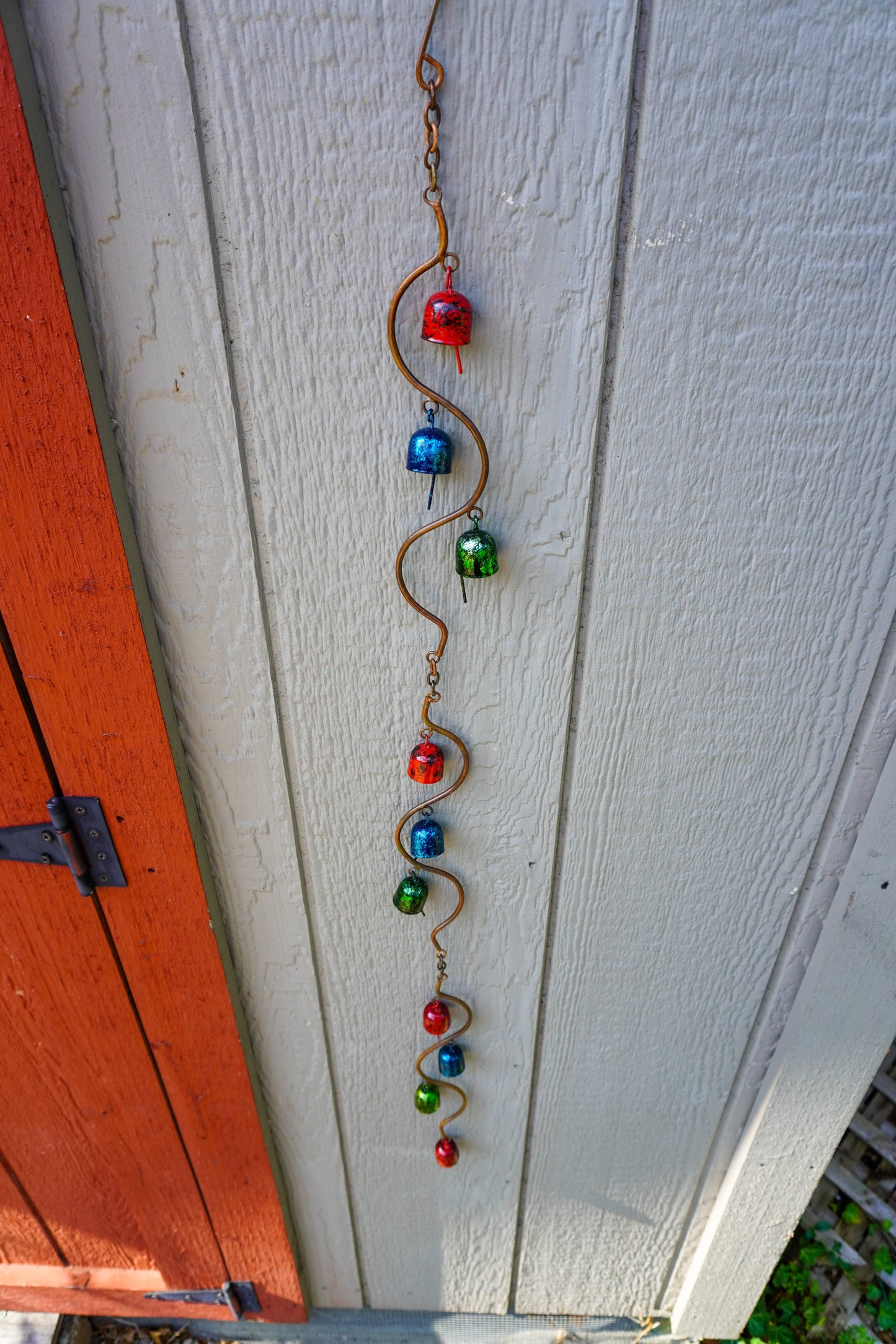 A vertical metal garden ornament with a wavy copper wire frame and colorful metal beads in red, blue, and green, hanging against a white painted wooden siding next to an orange-red door.