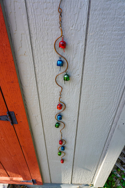 A vertical metal garden ornament with a wavy copper wire frame and colorful metal beads in red, blue, and green, hanging against a white painted wooden siding next to an orange-red door.