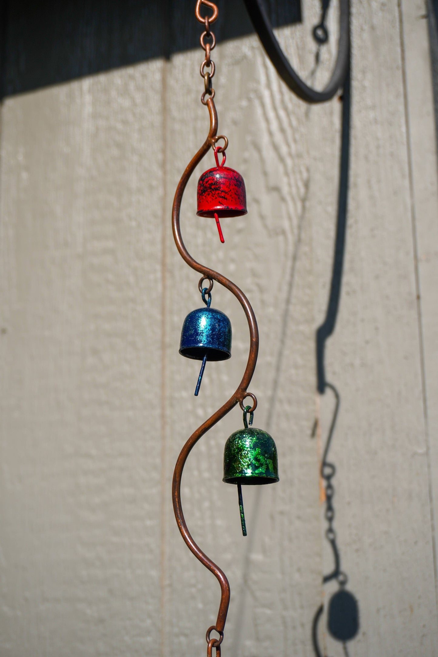 A copper-colored metal wind chime with three hanging bells in red, blue, and green, photographed against a light textured wall.