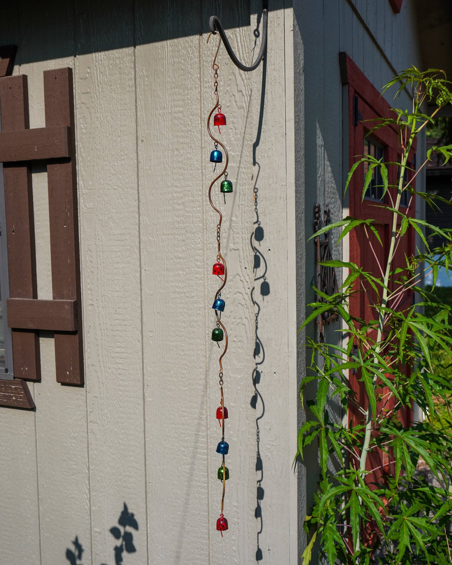 A decorative metal garden ornament hanging outdoors on a beige house wall, with green plants and a red window visible nearby.