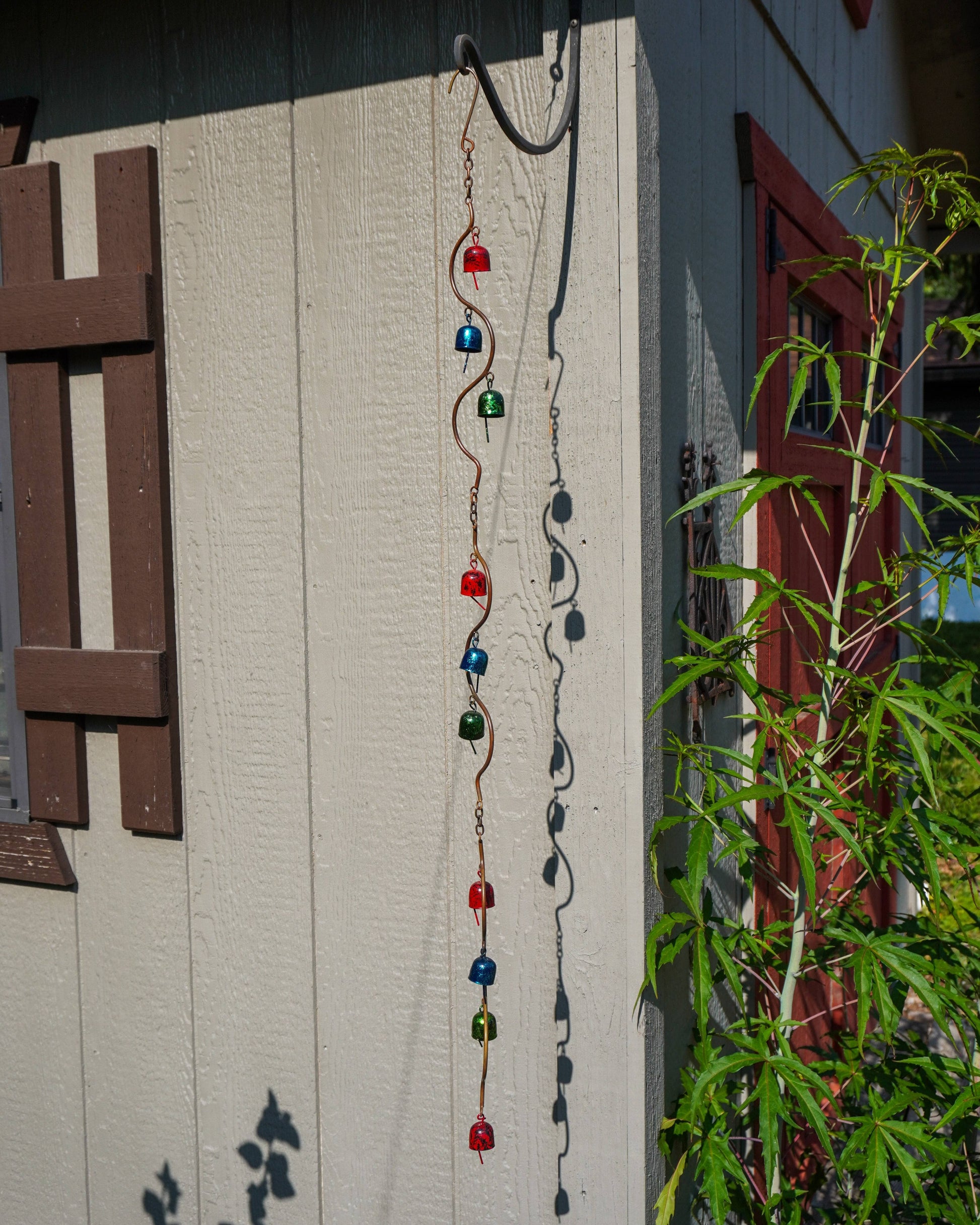 A decorative metal garden ornament hanging outdoors on a beige house wall, with green plants and a red window visible nearby.
