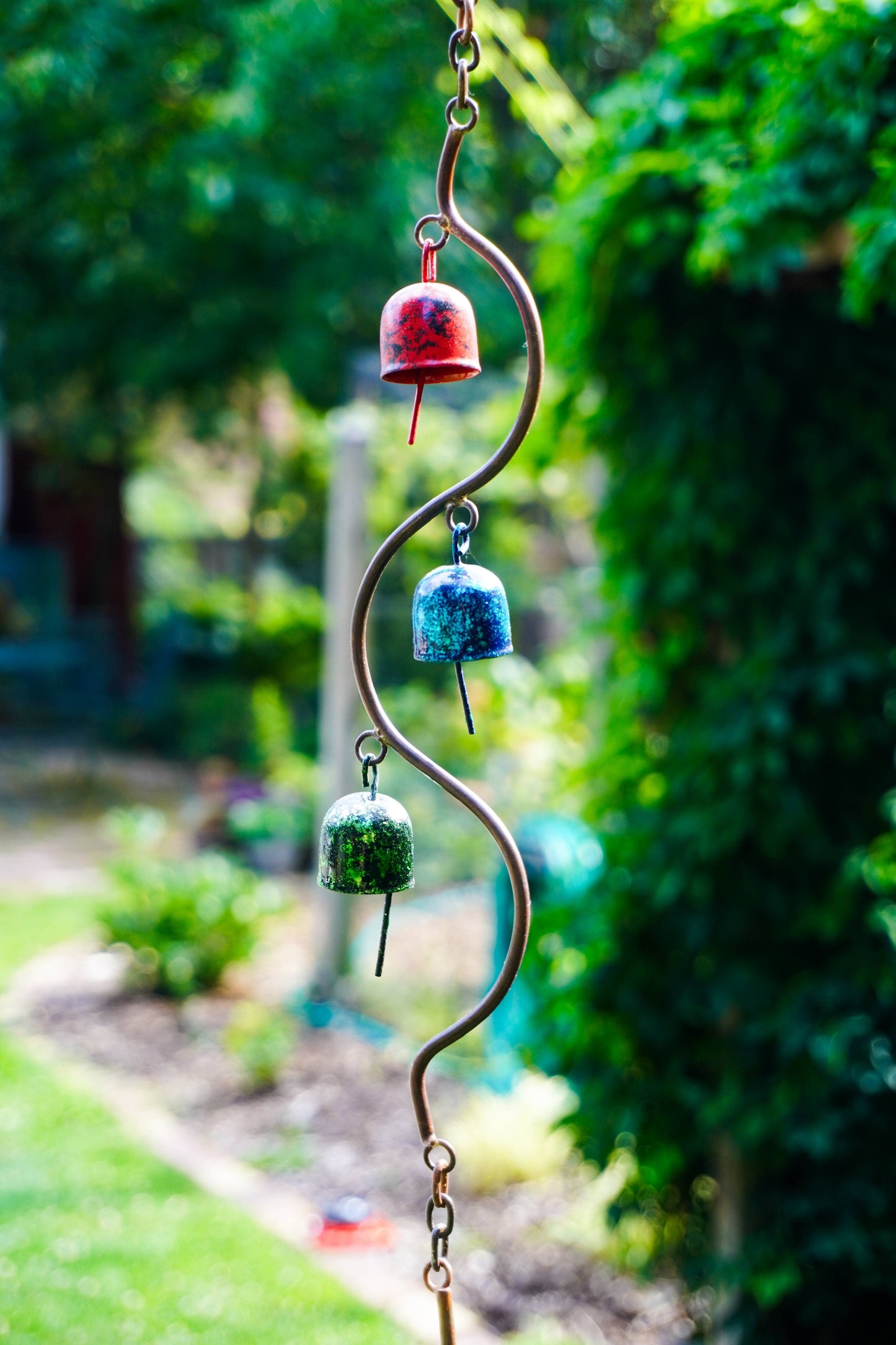 A hanging metal garden wind chime with three small bell-shaped ornaments in red, blue, and green, suspended from a curved metal rod in an outdoor garden setting.