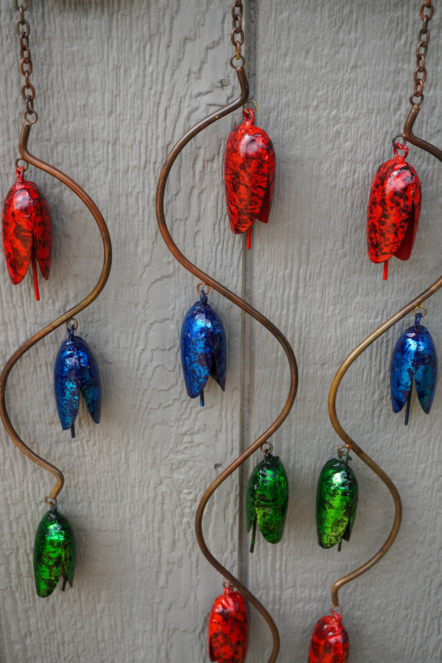 Three copper spiral wind chimes hanging against a light gray textured wall, each adorned with multiple colorful glass acorn charms in red, blue, and green.