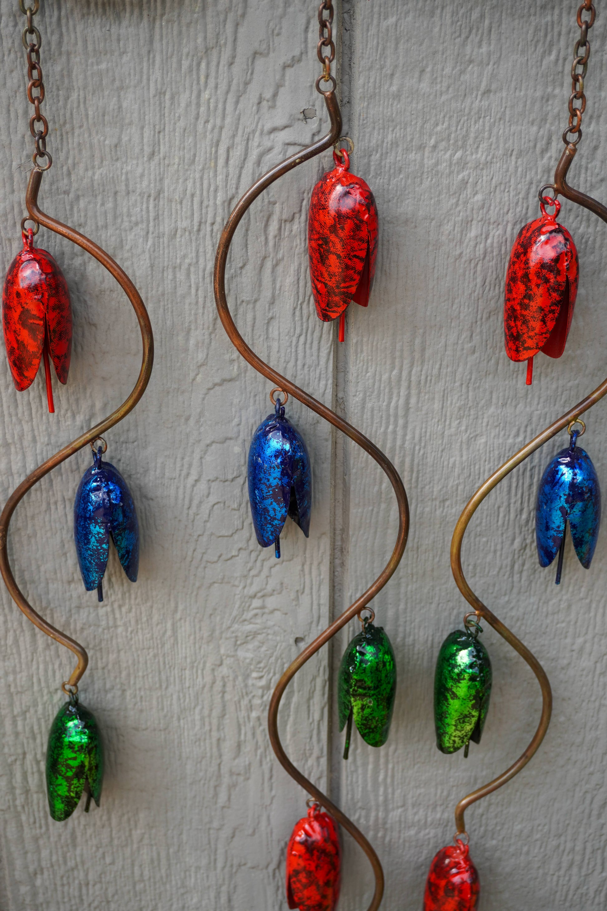 Three copper spiral wind chimes hanging against a light gray textured wall, each adorned with multiple colorful glass acorn charms in red, blue, and green.