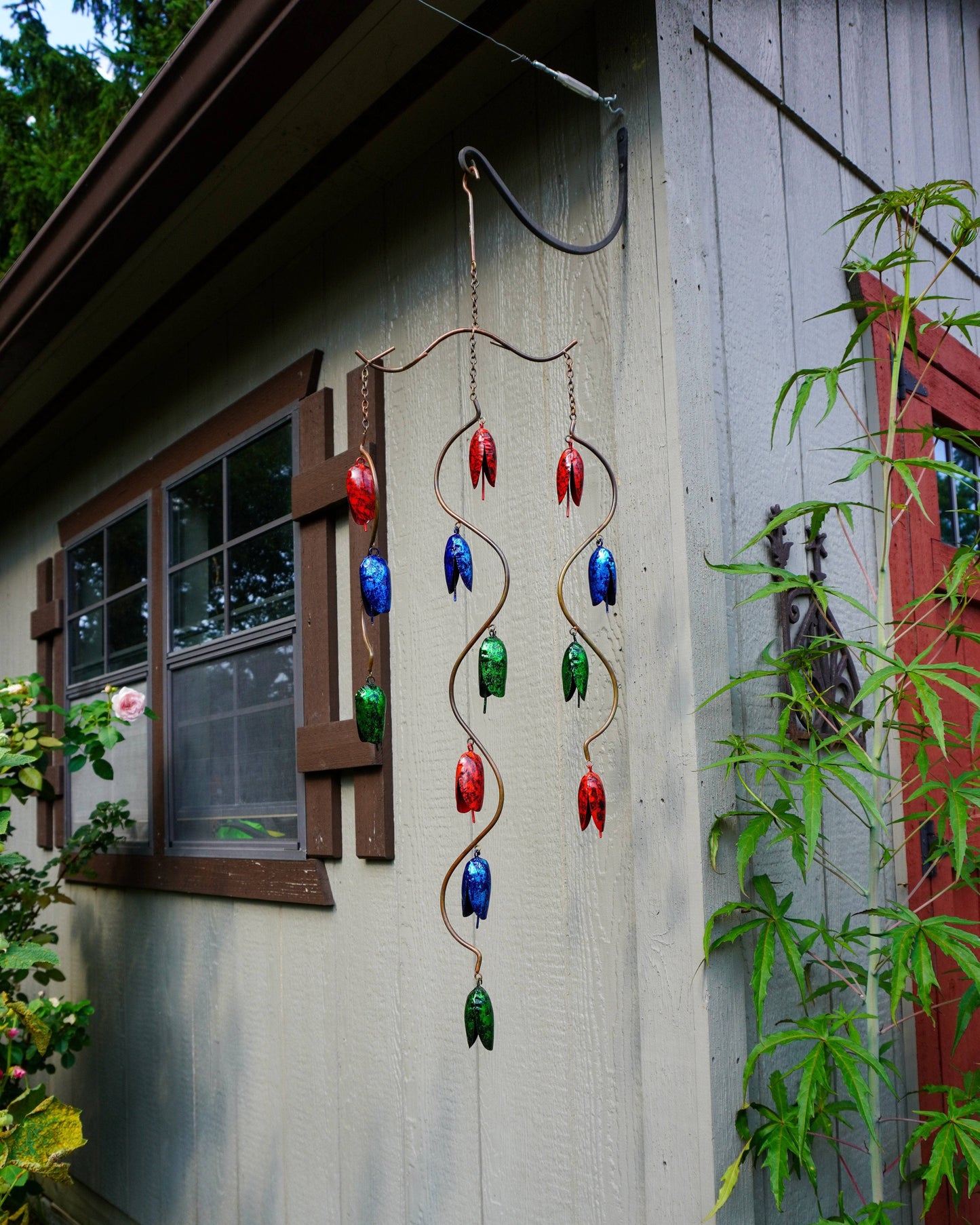 A hanging metal wind chime with red, blue, and green bell-shaped ornaments suspended from twisted wire, displayed outdoors against a gray house wall with plants and a window nearby.