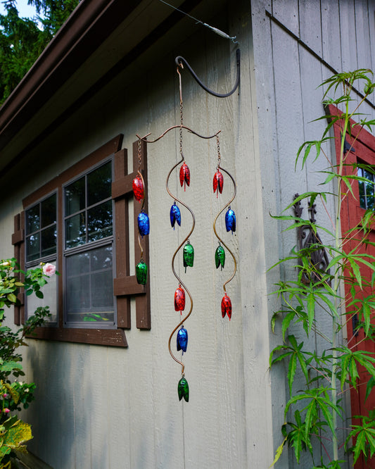A hanging metal wind chime with red, blue, and green bell-shaped ornaments suspended from twisted wire, displayed outdoors against a gray house wall with plants and a window nearby.