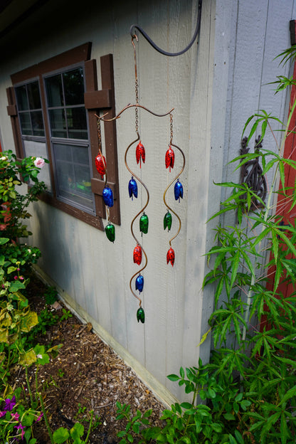 A copper wind spinner garden ornament with red, blue, and green glass tulip-like beads hangs on the exterior wall of a house next to green plants and mulch.