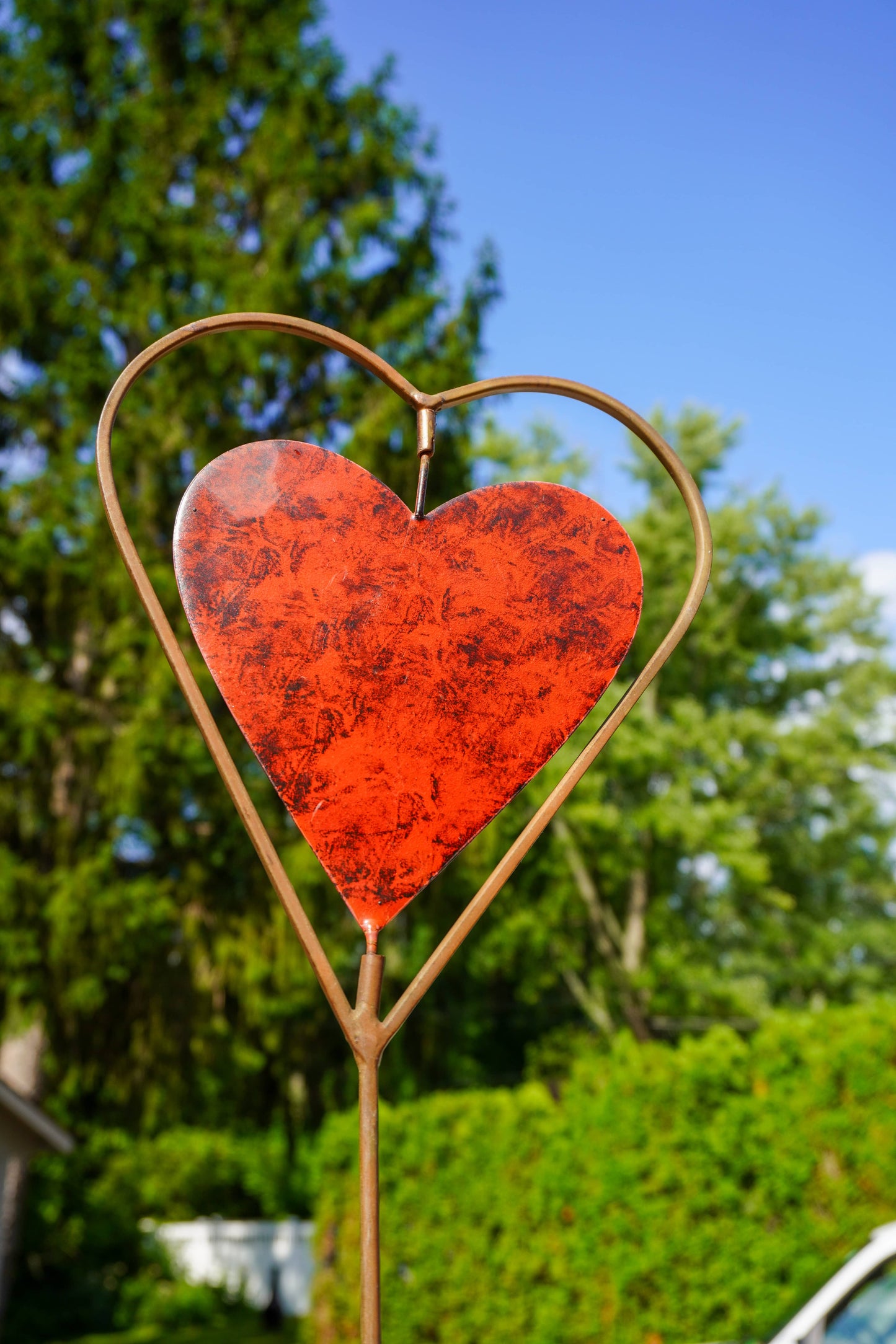 A heart-shaped metal garden stake with a textured red-orange glass heart insert, photographed outdoors against a background of green trees and blue sky.
