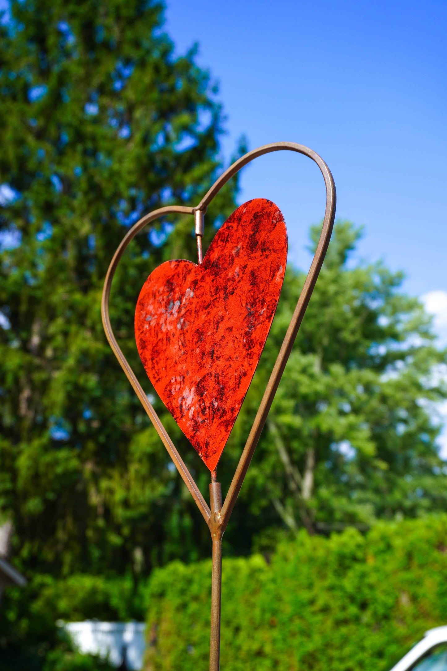 A decorative metal garden stake featuring a textured red heart in the center, set against a blurred background of green trees and blue sky.