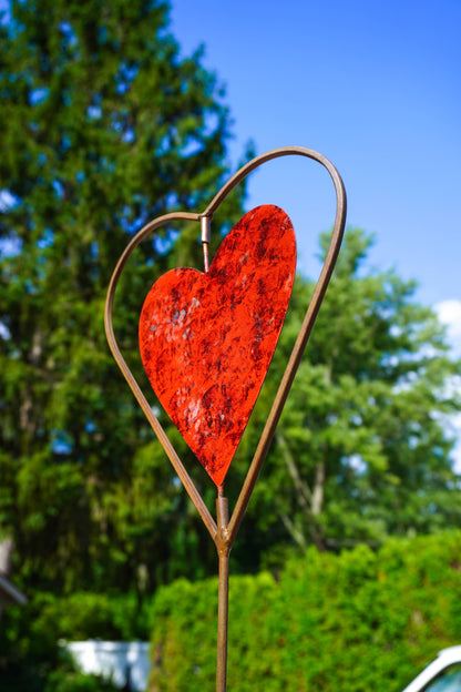 A decorative metal garden stake featuring a textured red heart in the center, set against a blurred background of green trees and blue sky.