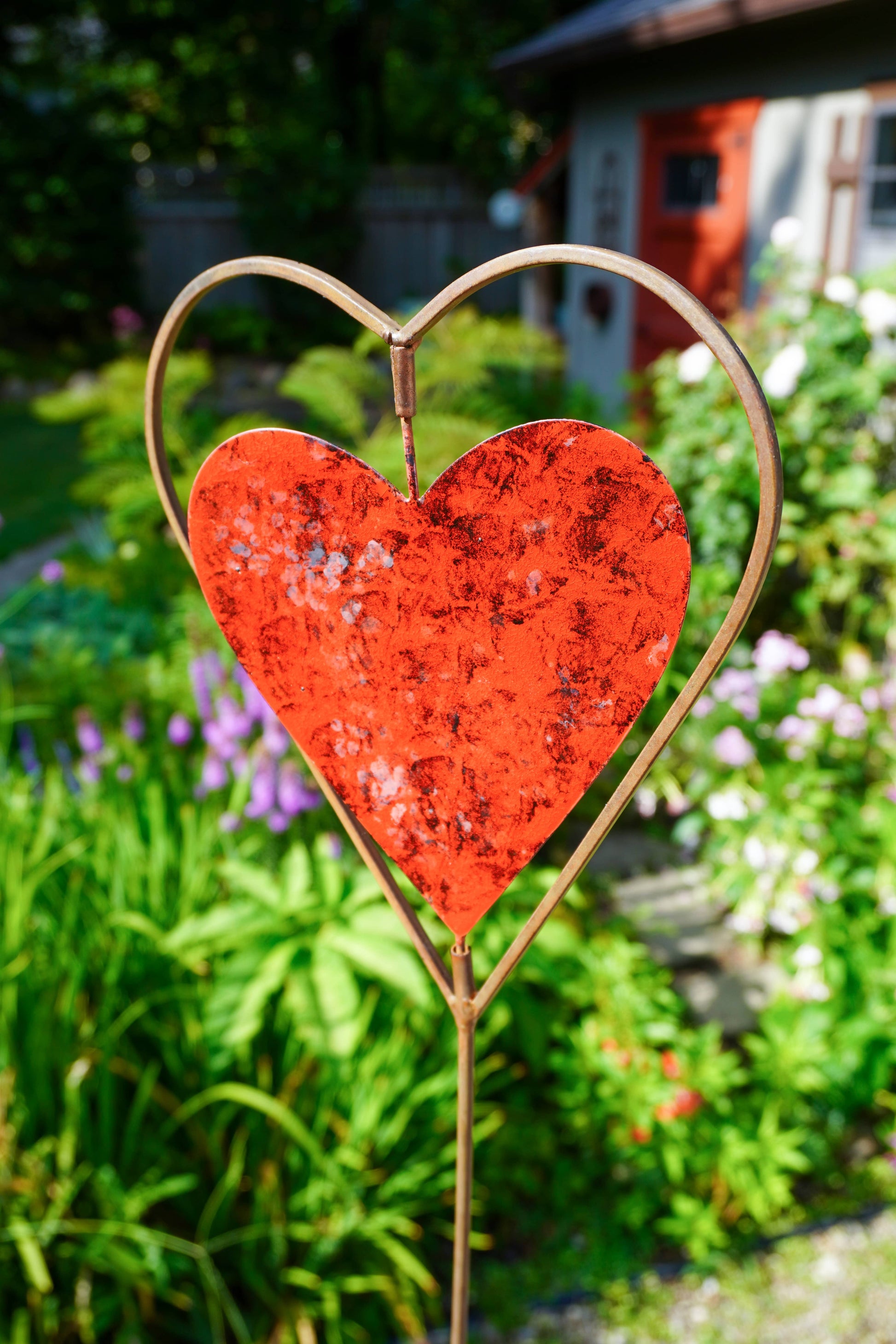 A rustic red heart-shaped metal garden stake with a gold-toned wire heart outline, set in a lush green garden.
