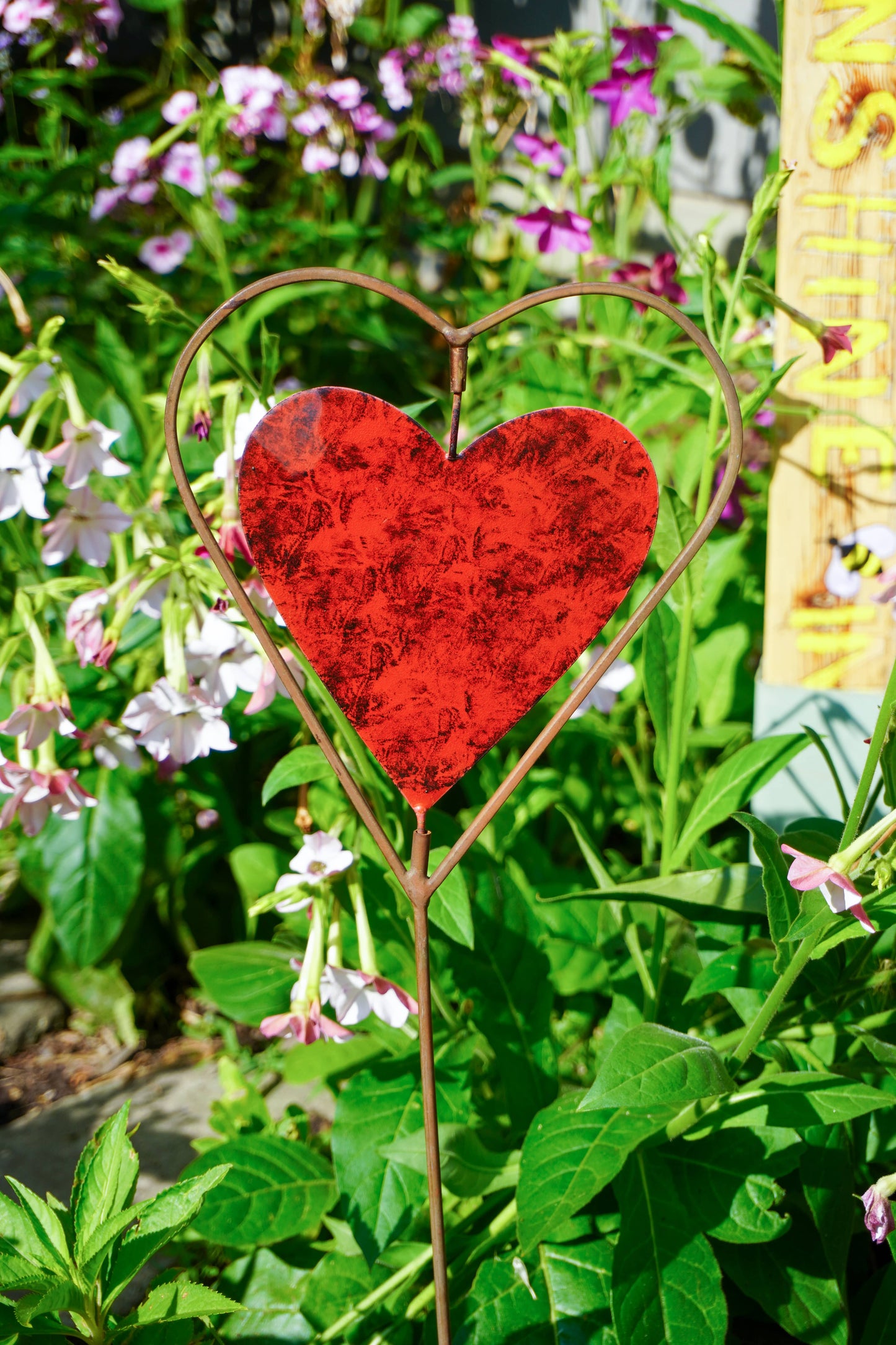A red heart-shaped metal garden stake with a rustic distressed texture, set in a lush garden with green foliage and pink flowers.